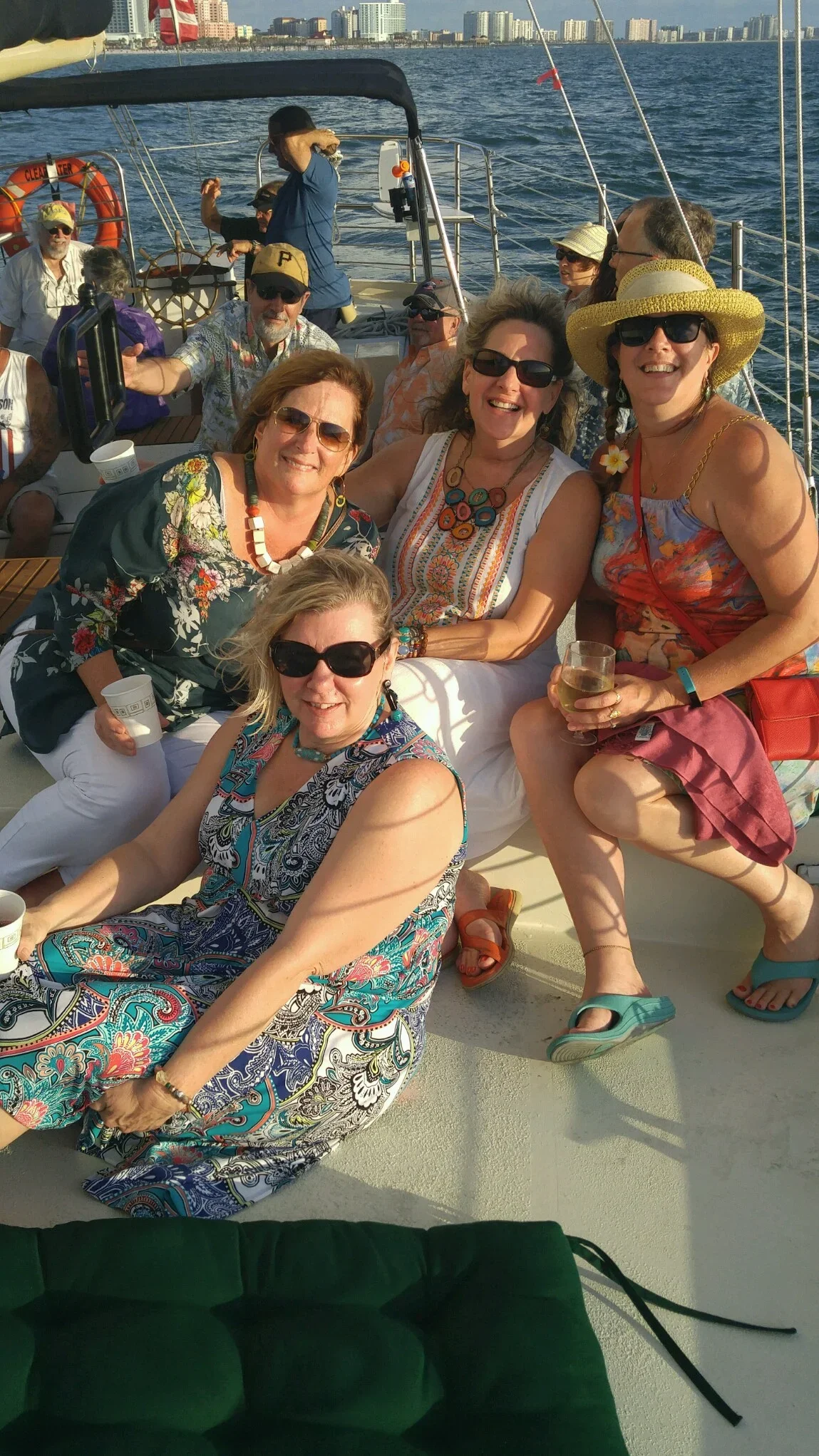 A group of people on a boat, enjoying a sunny day with city skyline in the background. Some are sitting and some standing, wearing casual summer clothes and sunglasses, holding drinks, smiling, and having a good time.