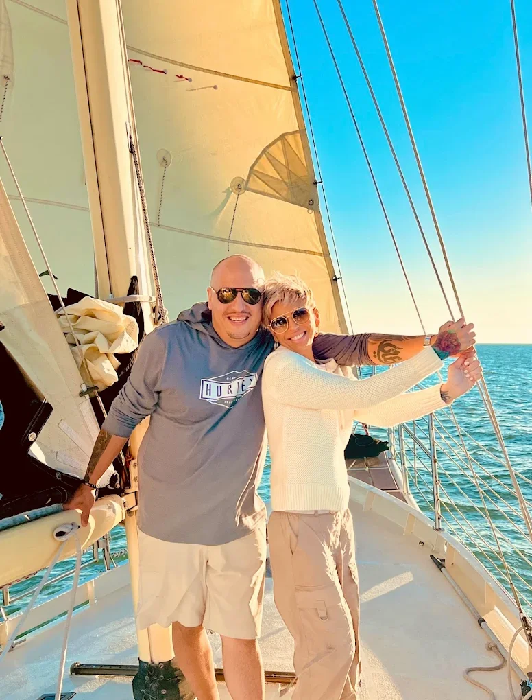 Two smiling people, a man and a woman, standing on a sailboat with blue ocean water in the background, sailing on a sunny day.