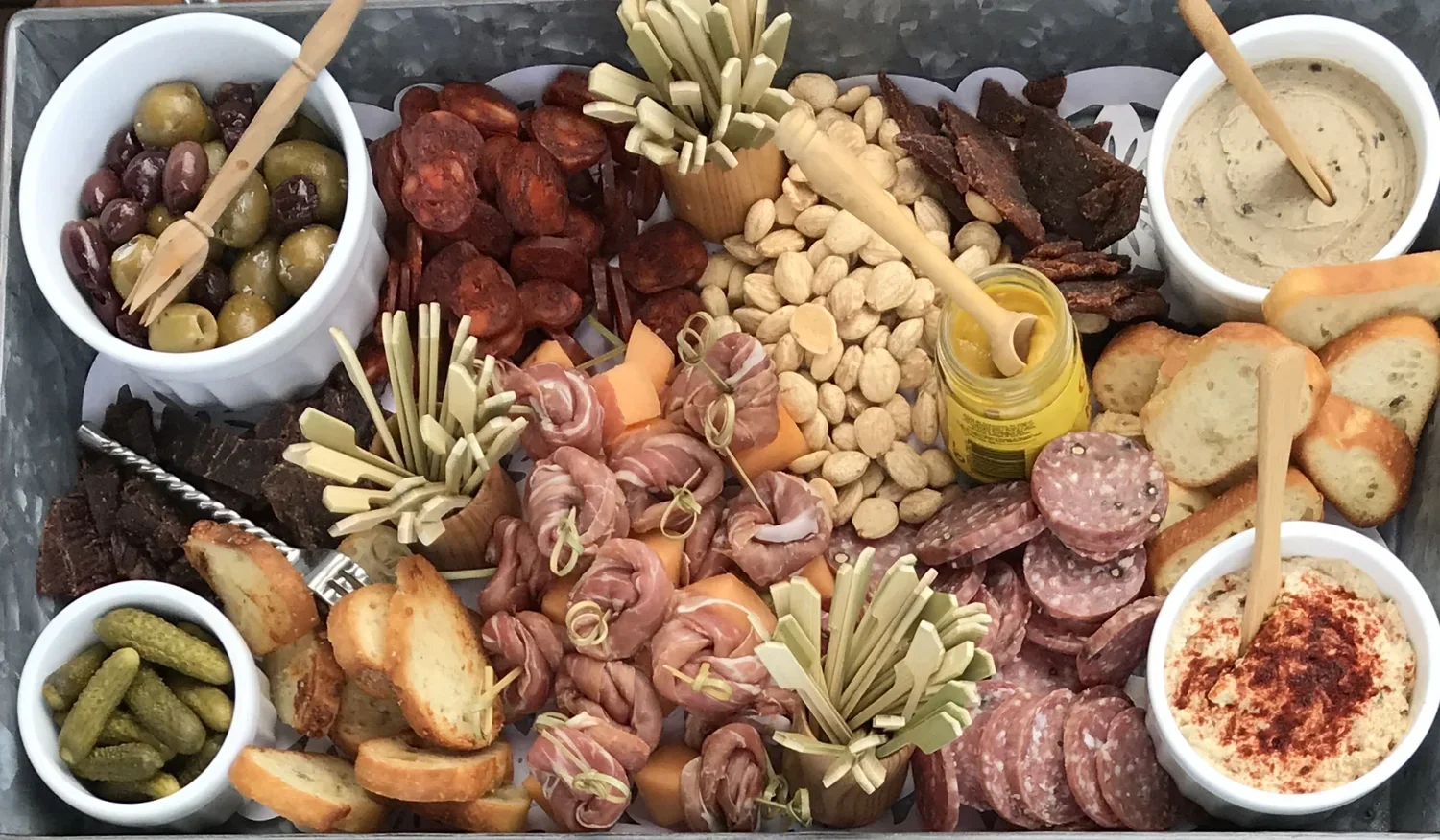 Charcuterie board with olives, cured meats, cheeses, bread, nuts, pickles, hummus, and condiments.