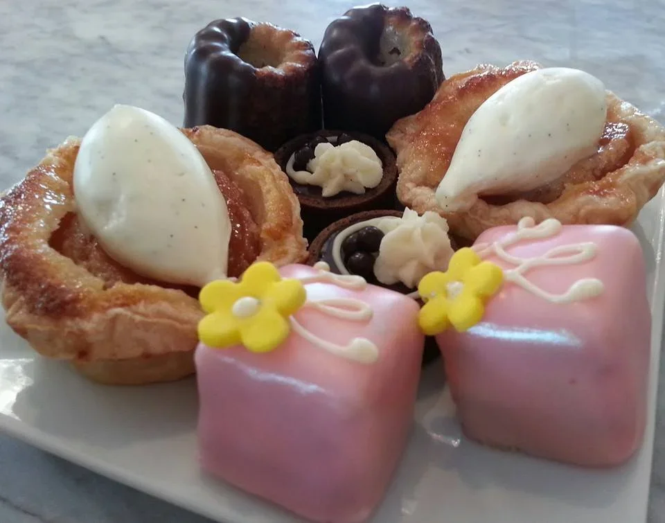 Assorted mini desserts including cannoli with white cream, chocolate cupcakes with white cream, and pink frosted cakes with yellow flower decorations.