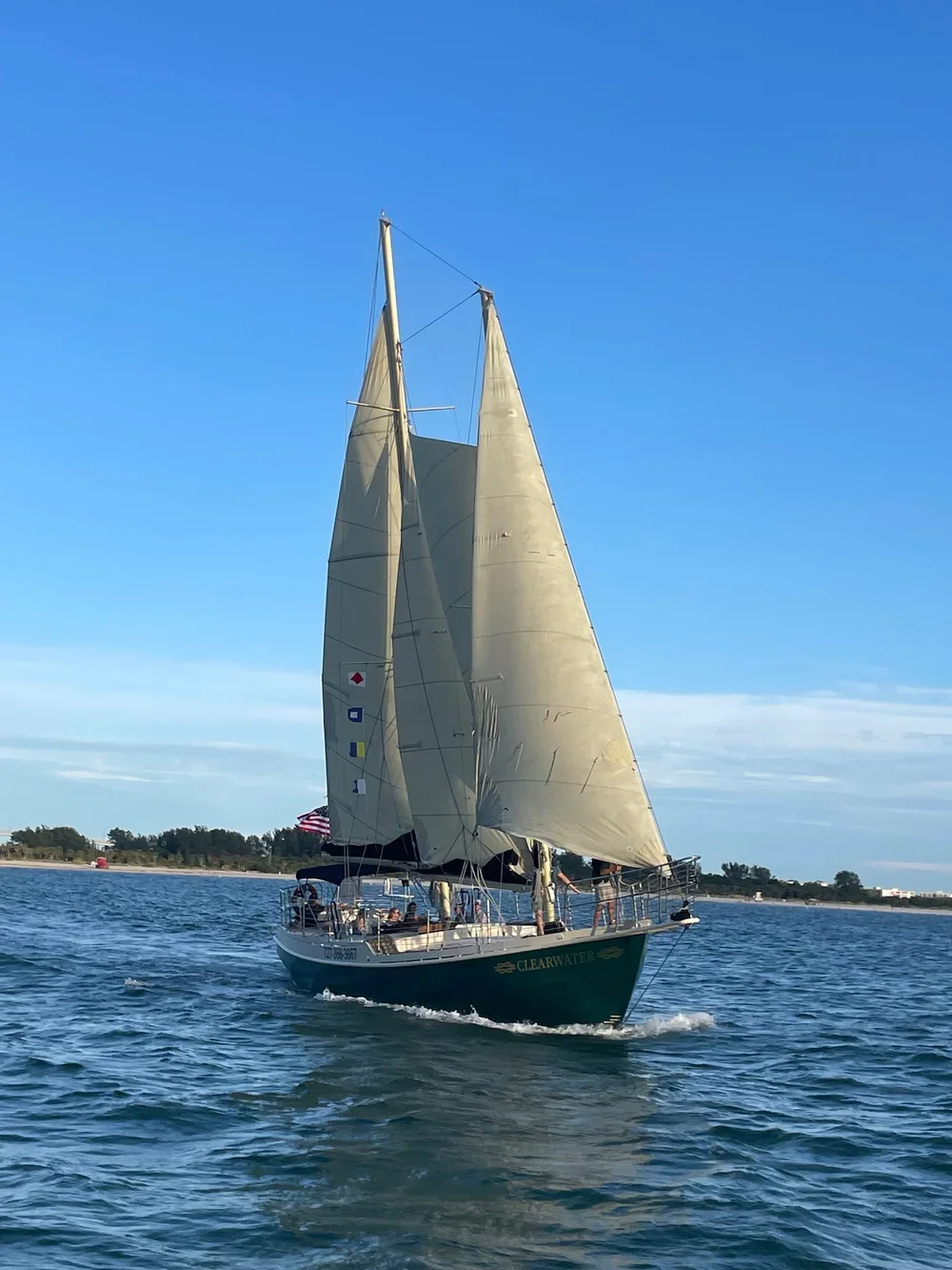 A sailboat with beige sails named Clearwater sailing on blue water with a land strip and trees in the background under a clear sky.