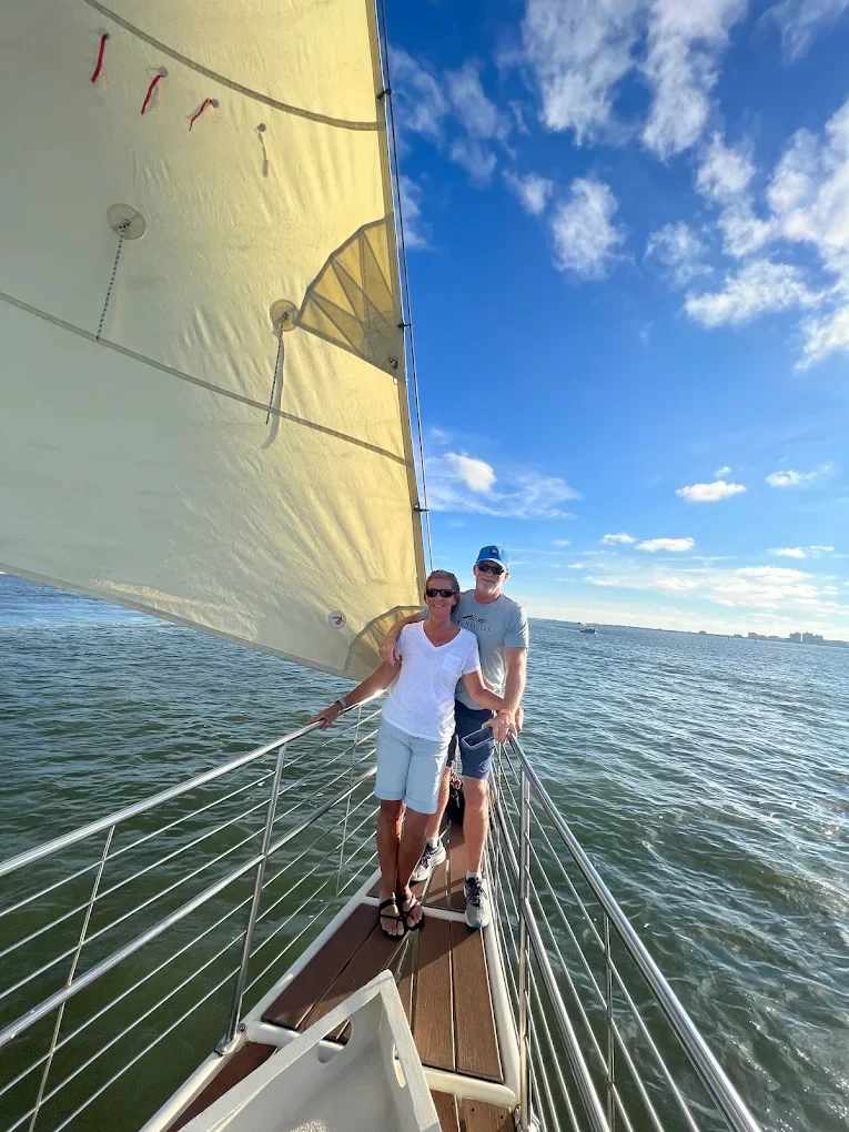 A couple standing on a sailboat's bow under a yellow sail, with one arm around each other, on a sunny day with a partly cloudy sky and a calm body of water.