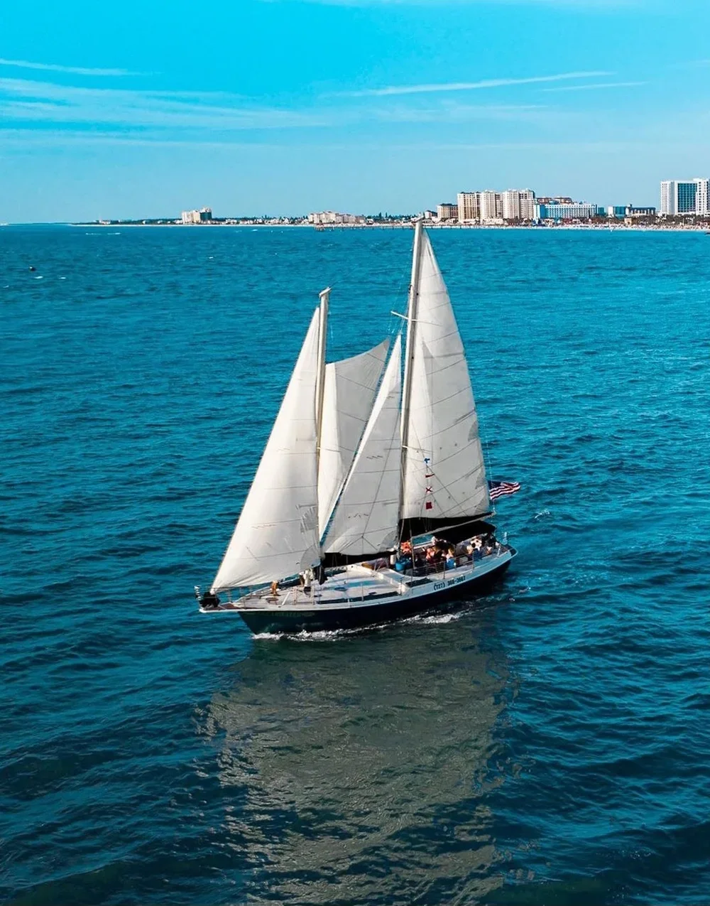 A sailboat with white sails sailing in blue ocean waters near a coastal city with high-rise buildings in the background.