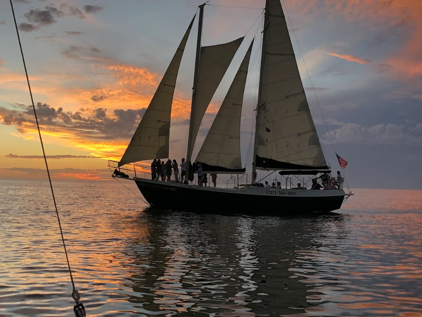 A sailboat with two masts sailing on calm water during sunset, with a colorful sky and scattered clouds in the background. A group of people is gathered on the deck, and an American flag is flying at the stern.