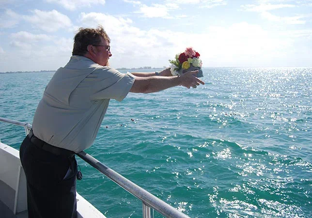 An older man on a boat holding a bouquet of flowers and an urn over the water, with the ocean in the background.