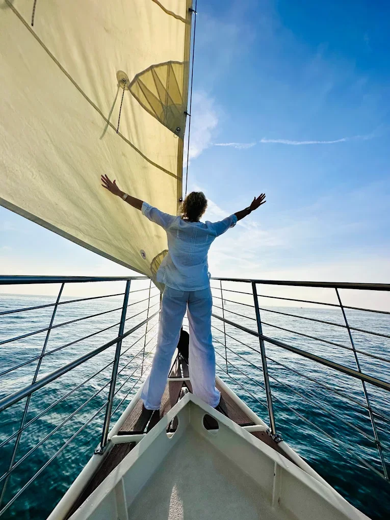 Person standing on a sailboat with arms outstretched, facing the water, under a blue sky with some clouds.