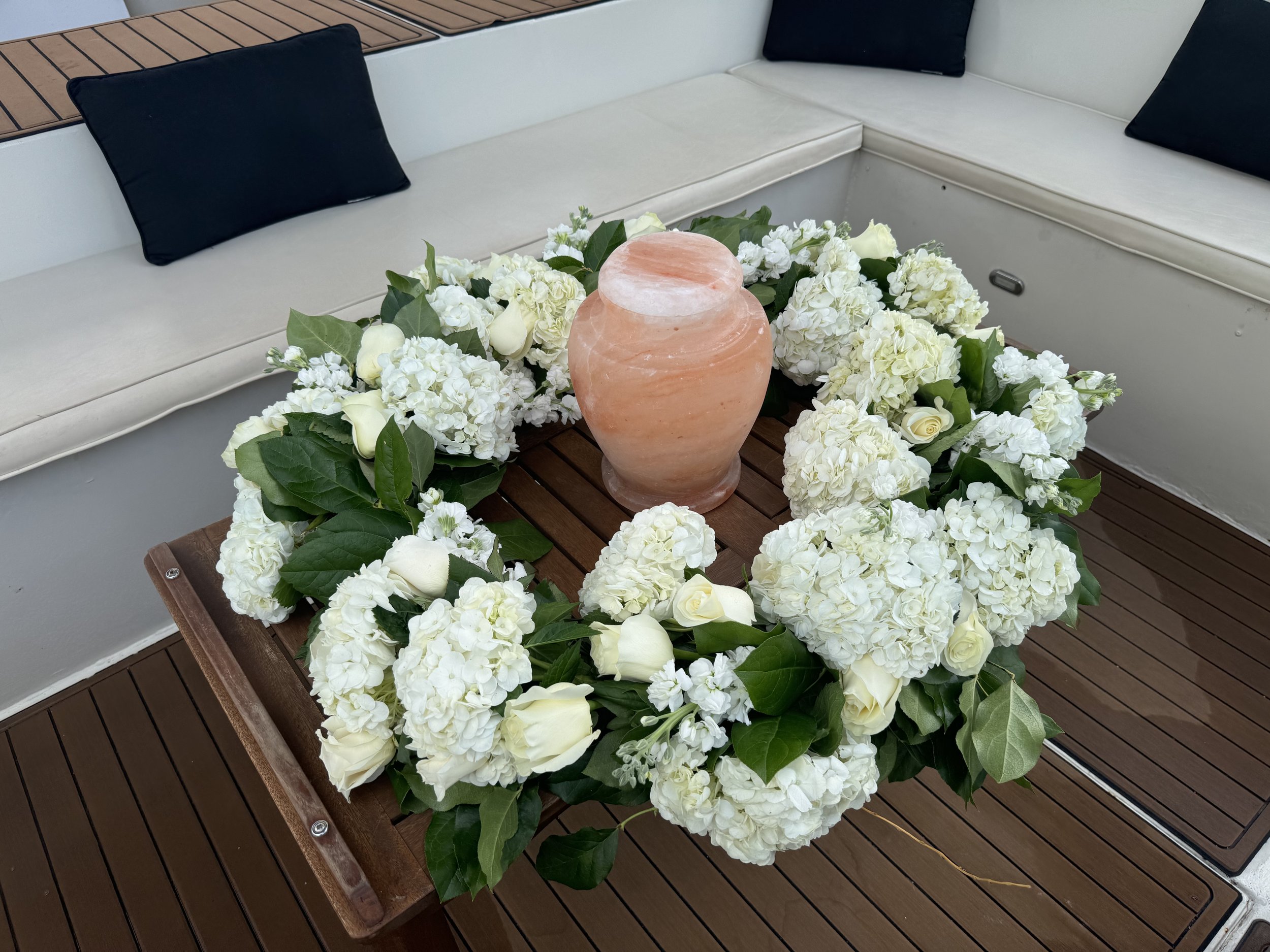 A floral arrangement with white hydrangeas, roses, and greenery surrounding a pink Himalayan salt urn on a wooden table.