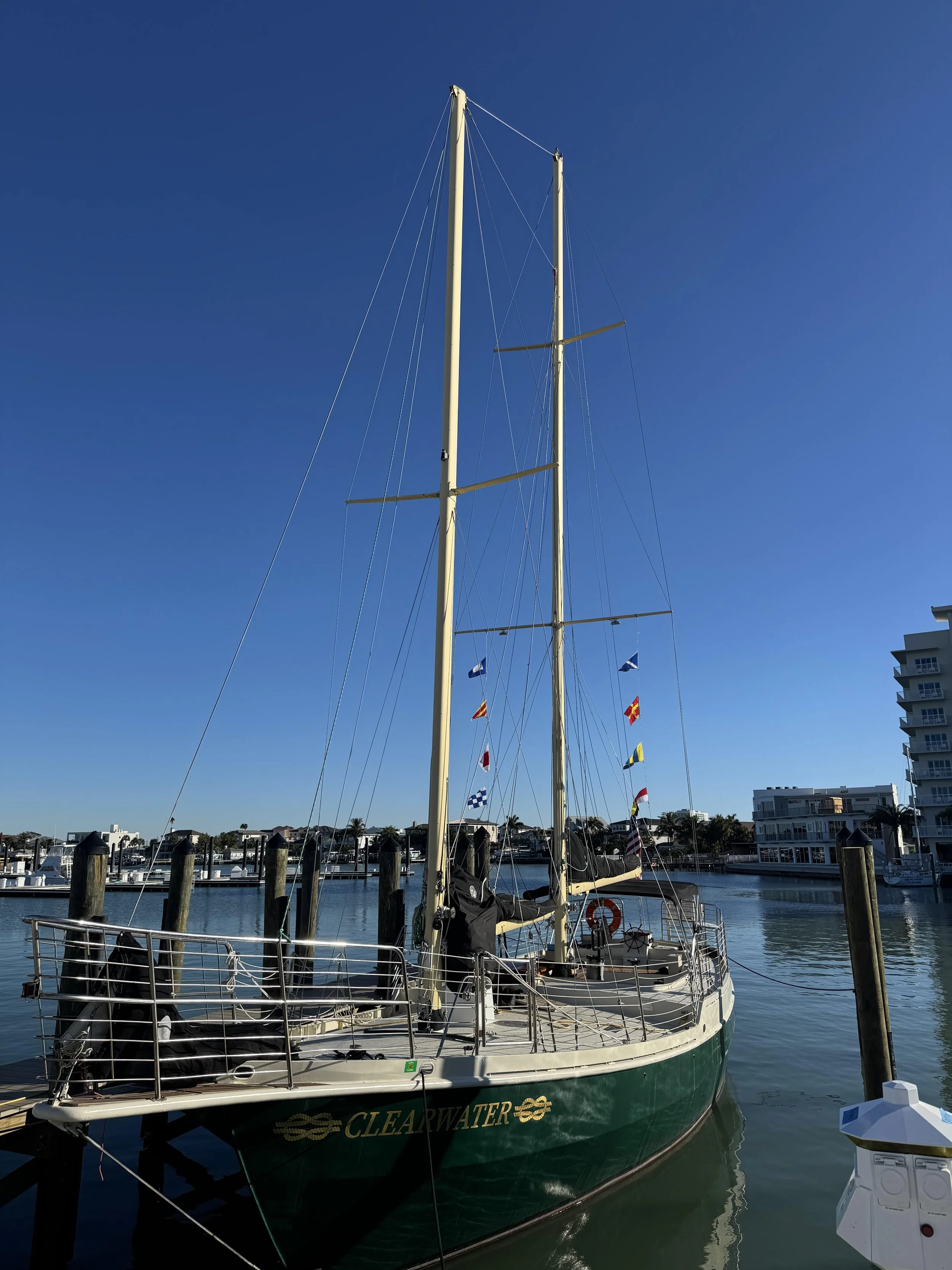 A sailboat named 'Clearwater' docked at a marina, with multiple flags hanging from its mast, calm water, and residential buildings in the background under a clear blue sky.
