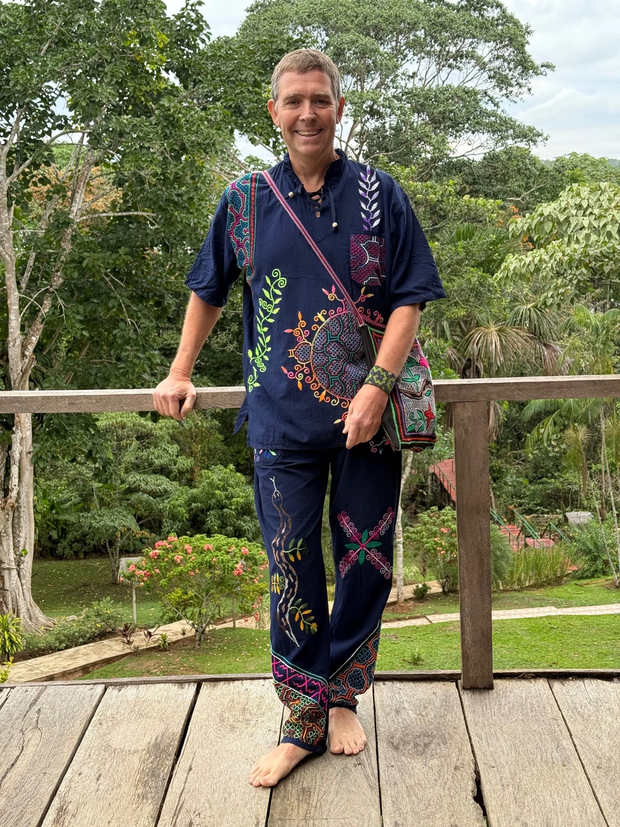 En person står på ett trädäck med gröna träd och buskar i bakgrunden. Personen är klädd i en blå broderad skjorta och byxor, bär en färgglad väska, och är barfota.
