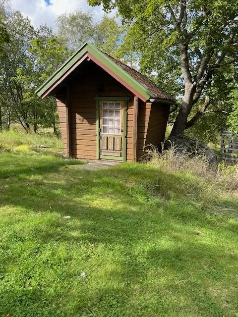 Litet trästugor i en grön trädgård med träd och gräs.