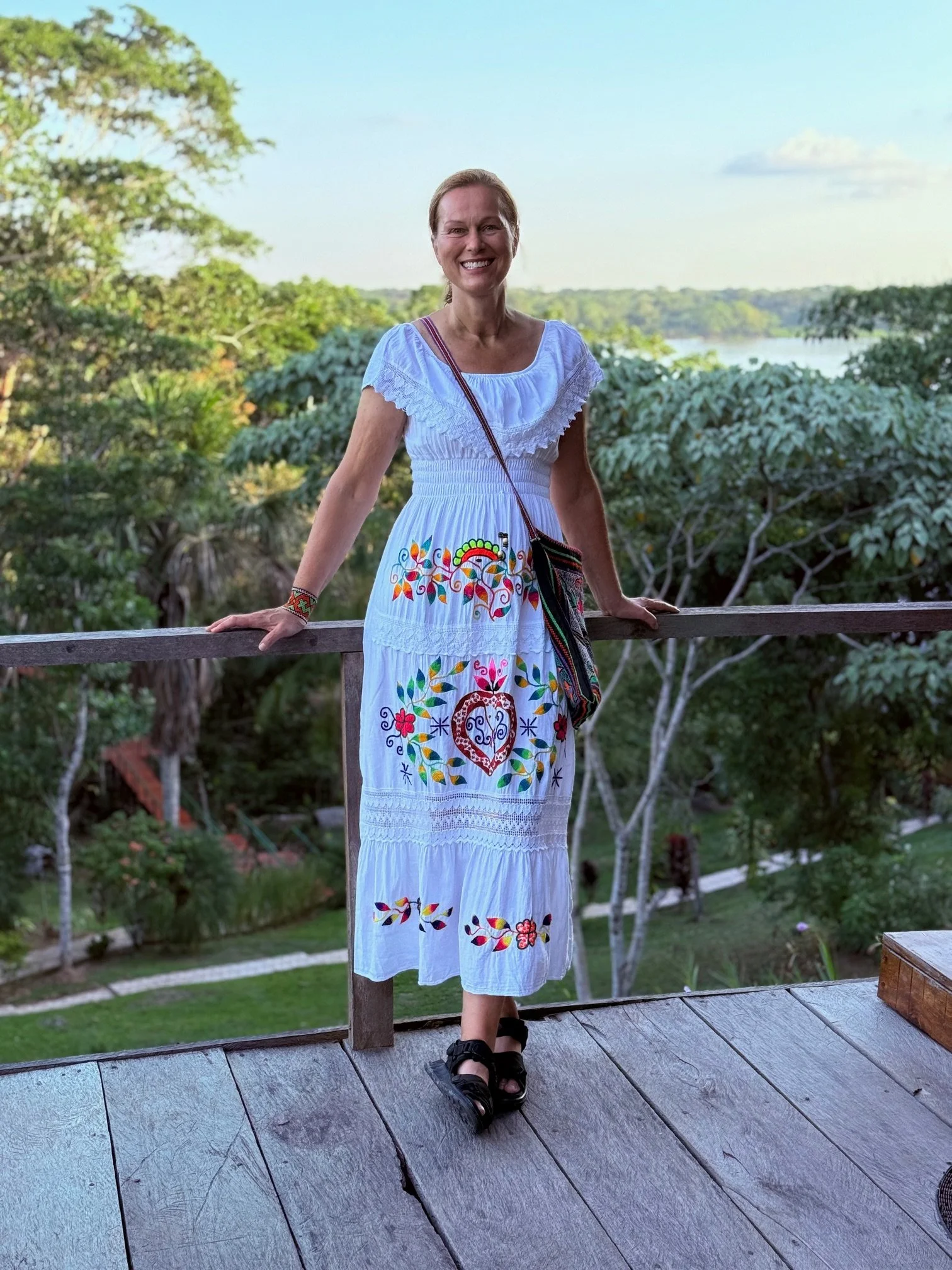 Kvinna i vit, handbroderad klänning står på en träplattform med utsikt över grönskande landskap och en flod i bakgrunden.