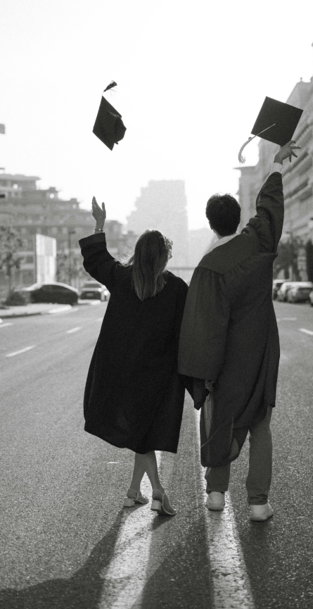 Two graduates in caps and gowns celebrate graduation on a city street with arms raised, throwing caps into the air.