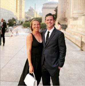 A man and woman dressed formally, standing outdoors in front of a grand building and a fountain, smiling for the photo.