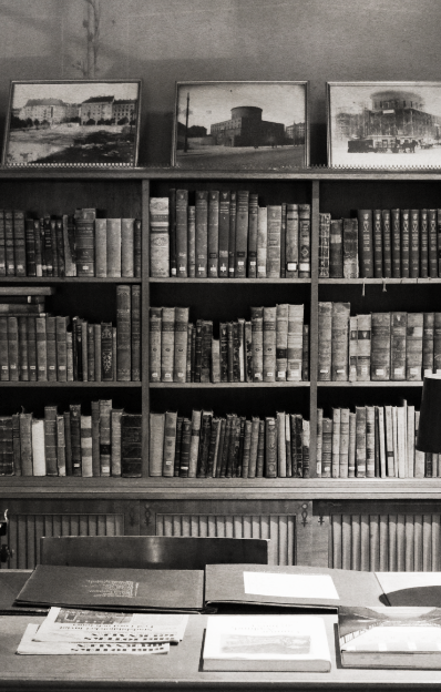 A black and white photo of a library with bookshelves filled with books, and framed pictures of buildings above the shelves. There is a desk with papers and a laptop in the foreground.