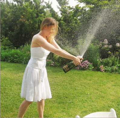 A woman in a white dress is outdoors opening a bottle of champagne, causing the cork and liquid to spray out.