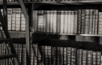Black and white photo of old books on wooden shelves, some books are stacked horizontally, a ladder leans against the shelf.