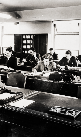Black and white photo of women working at desks in an office, with bookshelves and windows in the background.