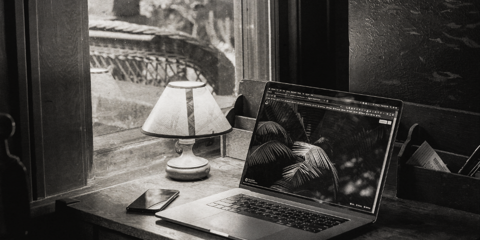 A laptop and smartphone on a wooden desk near a window with a lamp, inside a cozy room.