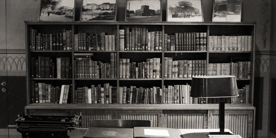 Black and white photo of a library with a bookshelf filled with books, a desk with papers, a typewriter, and a table lamp.