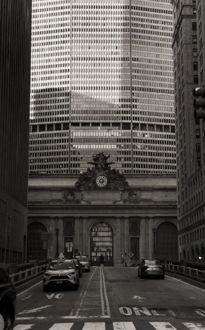 Black and white photo of a city street with tall skyscrapers, cars parked on each side, and a historic building with a clock and sculptures in the middle background.