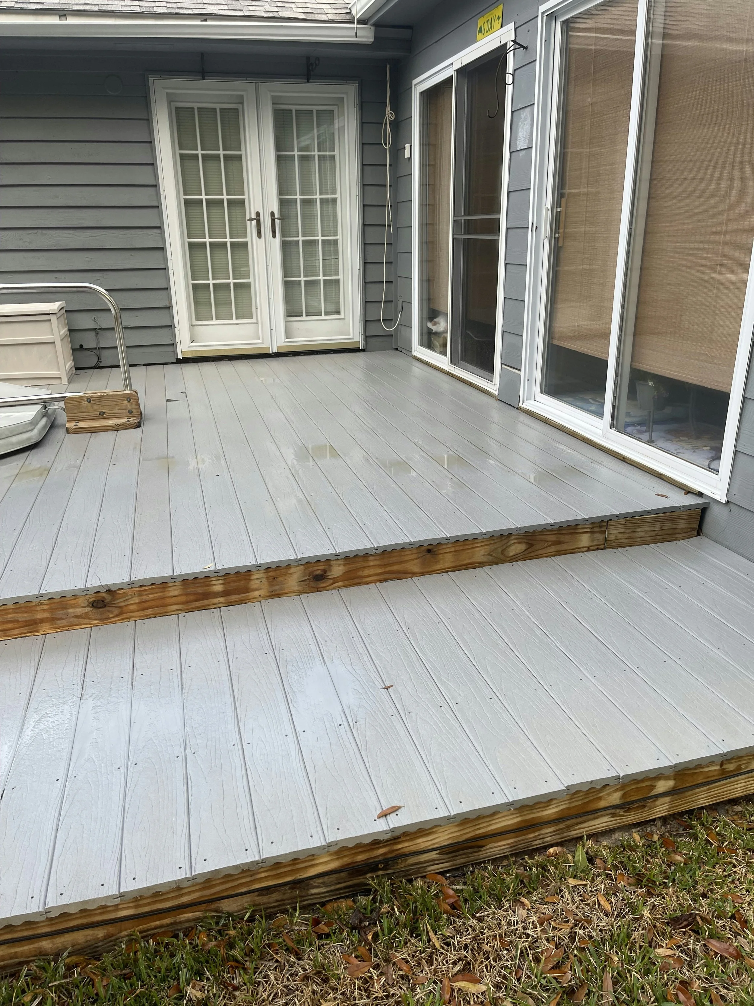Newly installed light gray porch decking with a small step, attached to a house with gray siding, double glass doors with white frames, and sliding screens.