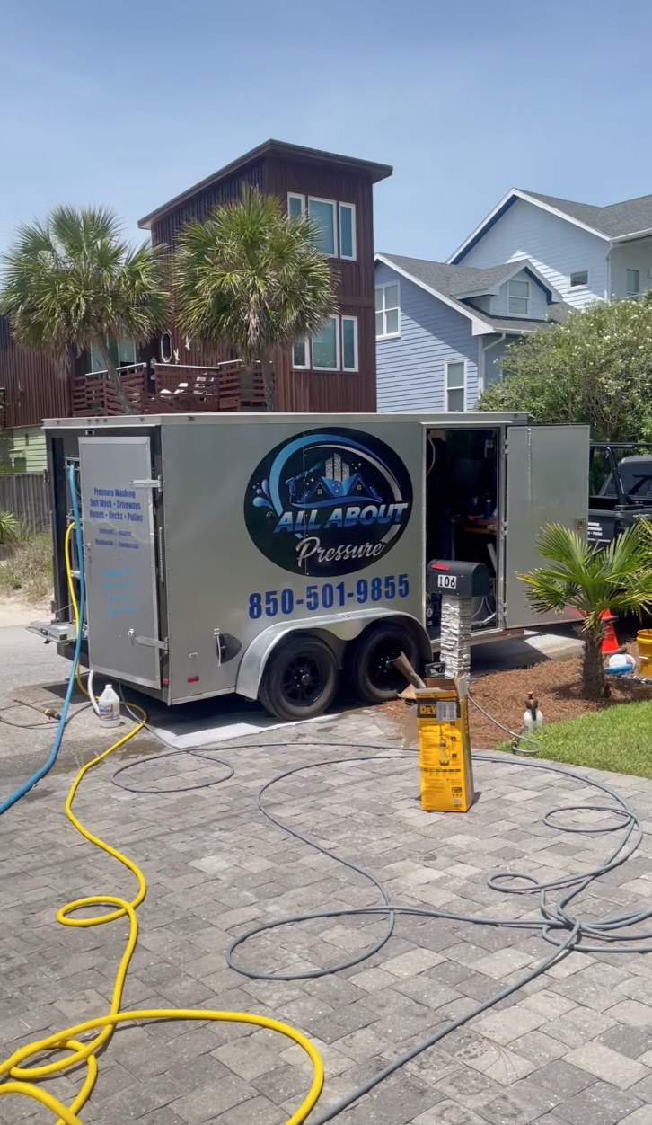 Pressure washing trailer with hoses on a brick driveway, surrounded by plants and residential houses.