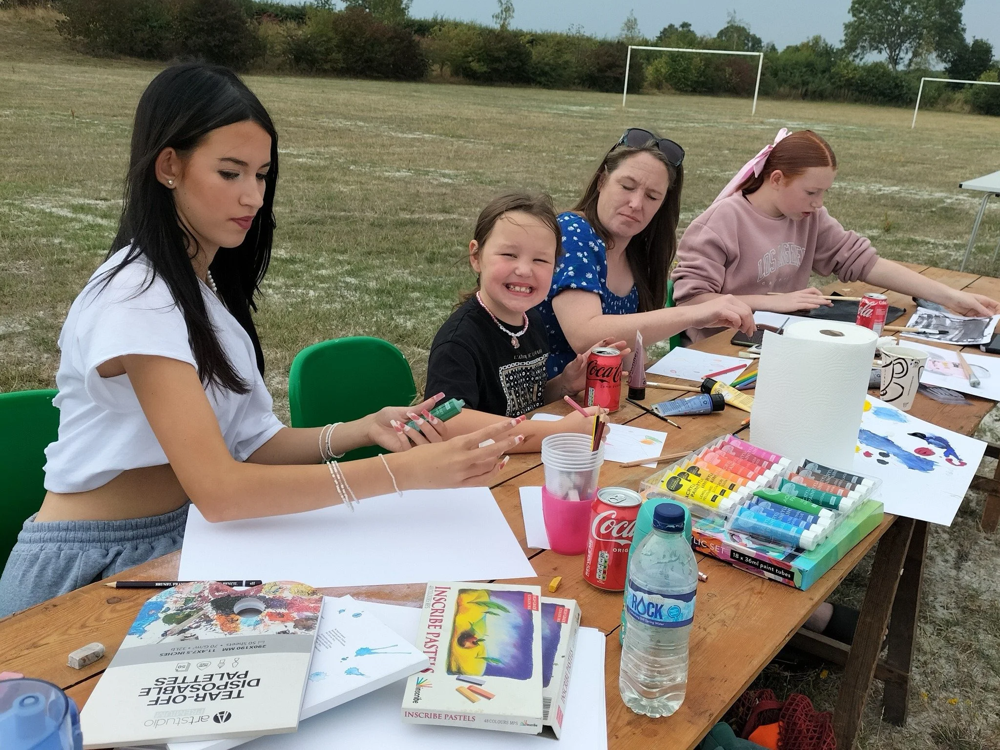 Four people sitting at a wooden outdoor table, engaging in arts and crafts with paints, colored pencils, and paper. The table holds various art supplies, soda cans, a water bottle, and books. A grassy field with soccer goals and trees is in the backg