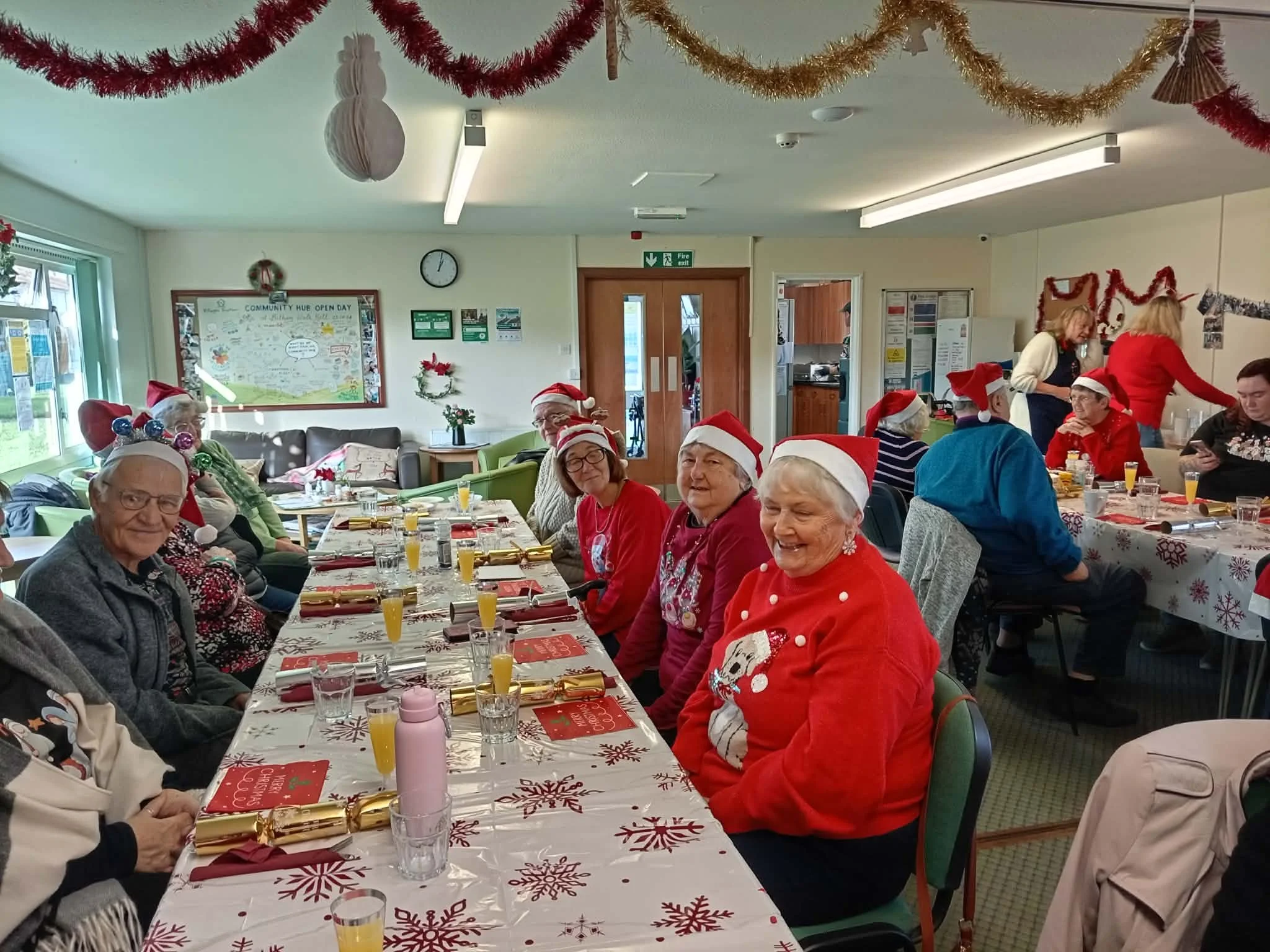 Elderly people wearing Christmas hats and sweaters sitting at a decorated holiday table, with Christmas tinsel and ornaments in a community room.