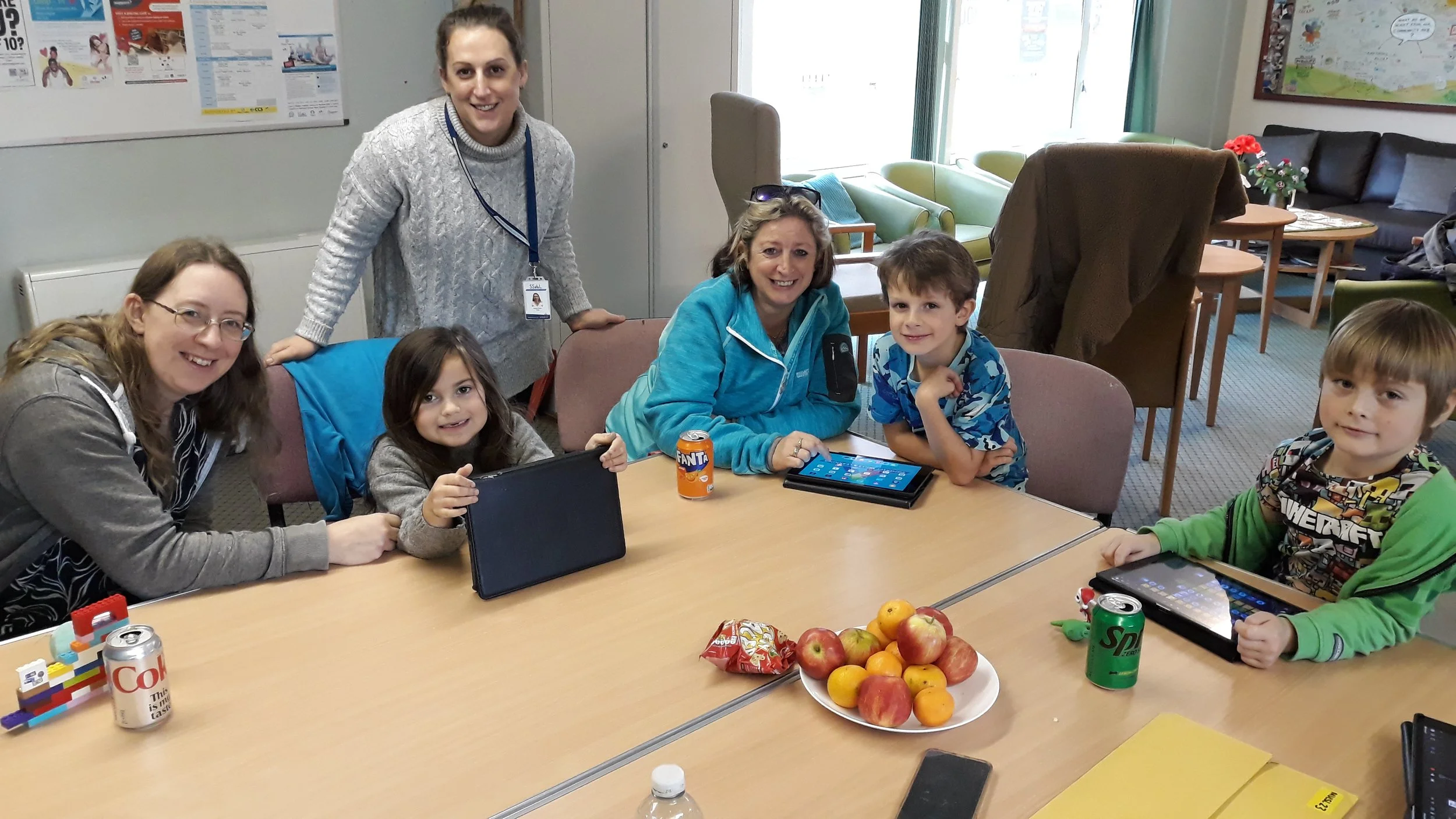 Two men sitting in armchairs engaged in a conversation indoors, with floor-to-ceiling vertical blinds/windows behind them. The man on the left has gray hair, wears a black hoodie, and has his chin resting on his hand. The man on the right has long dark hair, glasses, and is wearing a black T-shirt with patches that read "CAPTAIN" and "TALK CLUB." There is a table with flowers, a notepad, and some snacks in front of them.