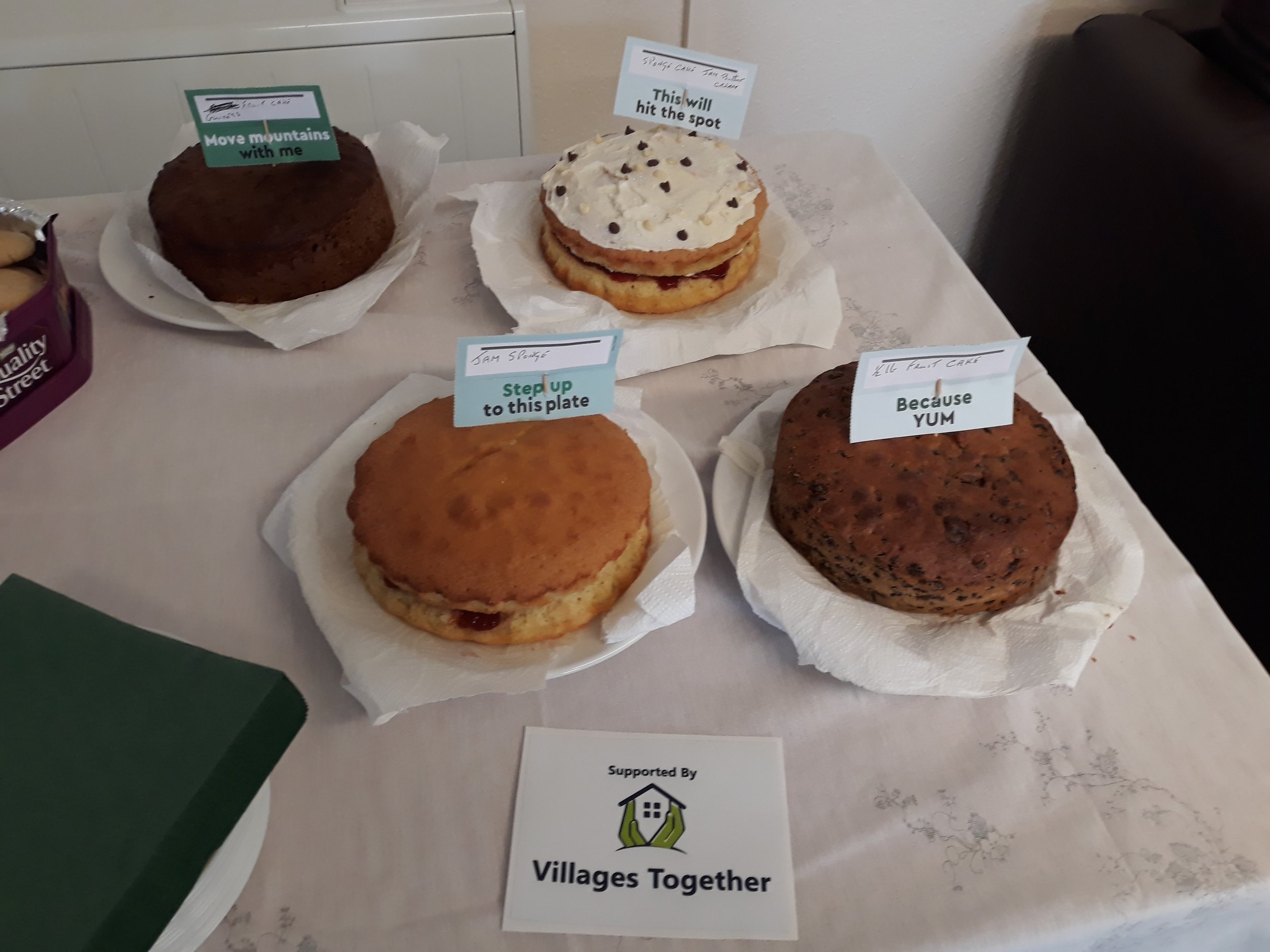 A display of four different cakes on a table with labels. The cakes include a chocolate cake labeled 'Move mountains with me,' a layered cake with cream and chocolate chips labeled 'This will hit the spot,' a sponge cake labeled 'Step up to this plat