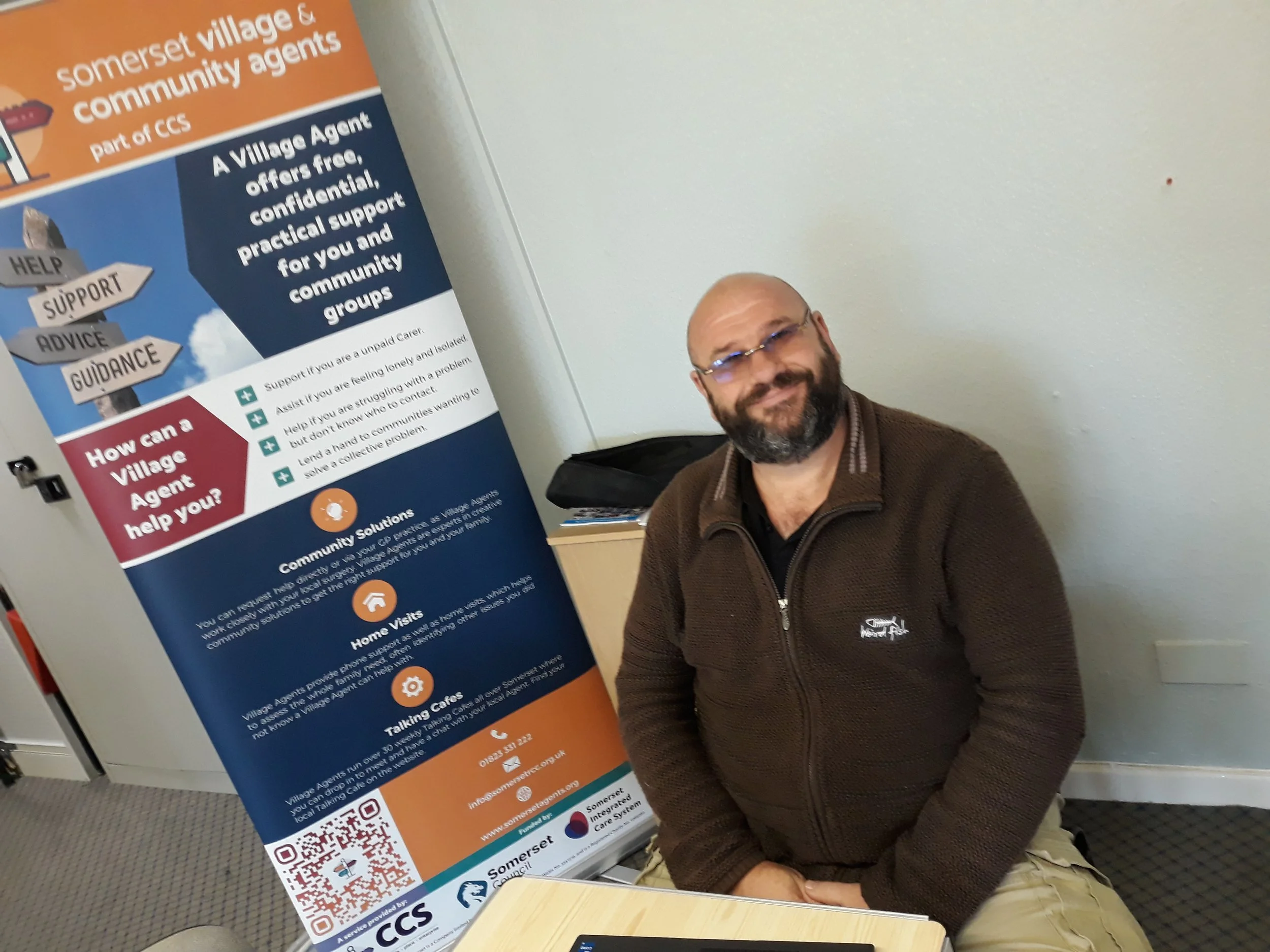Man with glasses and beard sitting next to a Somerset Village and community agents informational poster.
