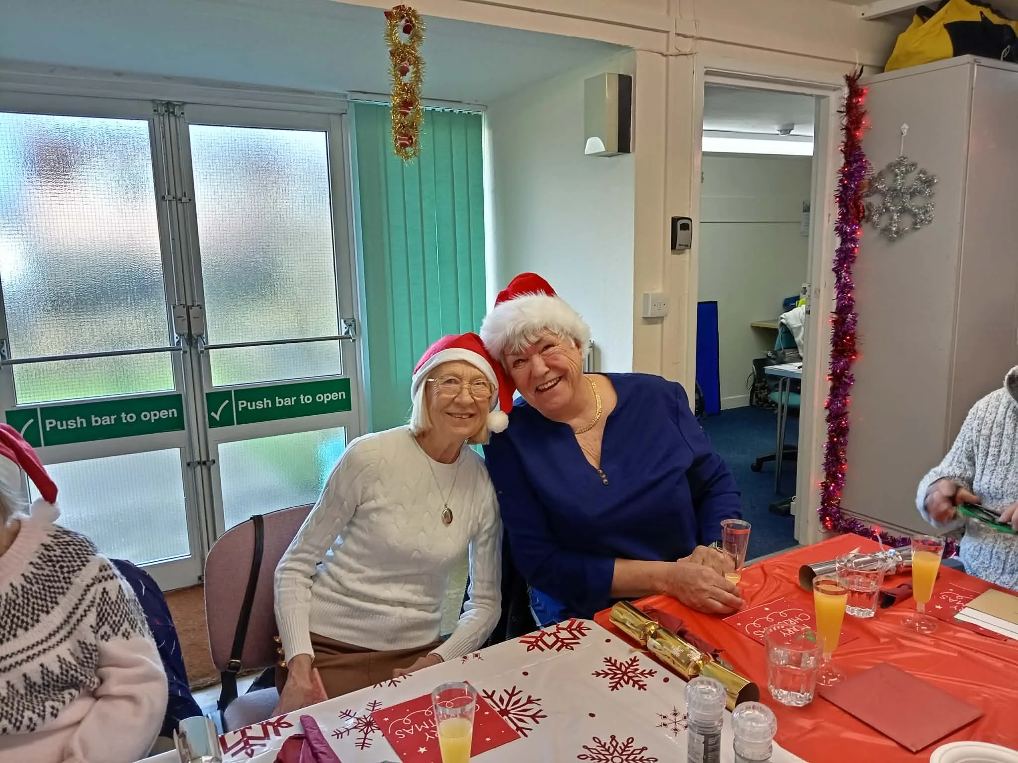 Two elderly women wearing Santa hats smiling at the camera during a Christmas celebration, sitting at a decorated table with drinks and gifts.