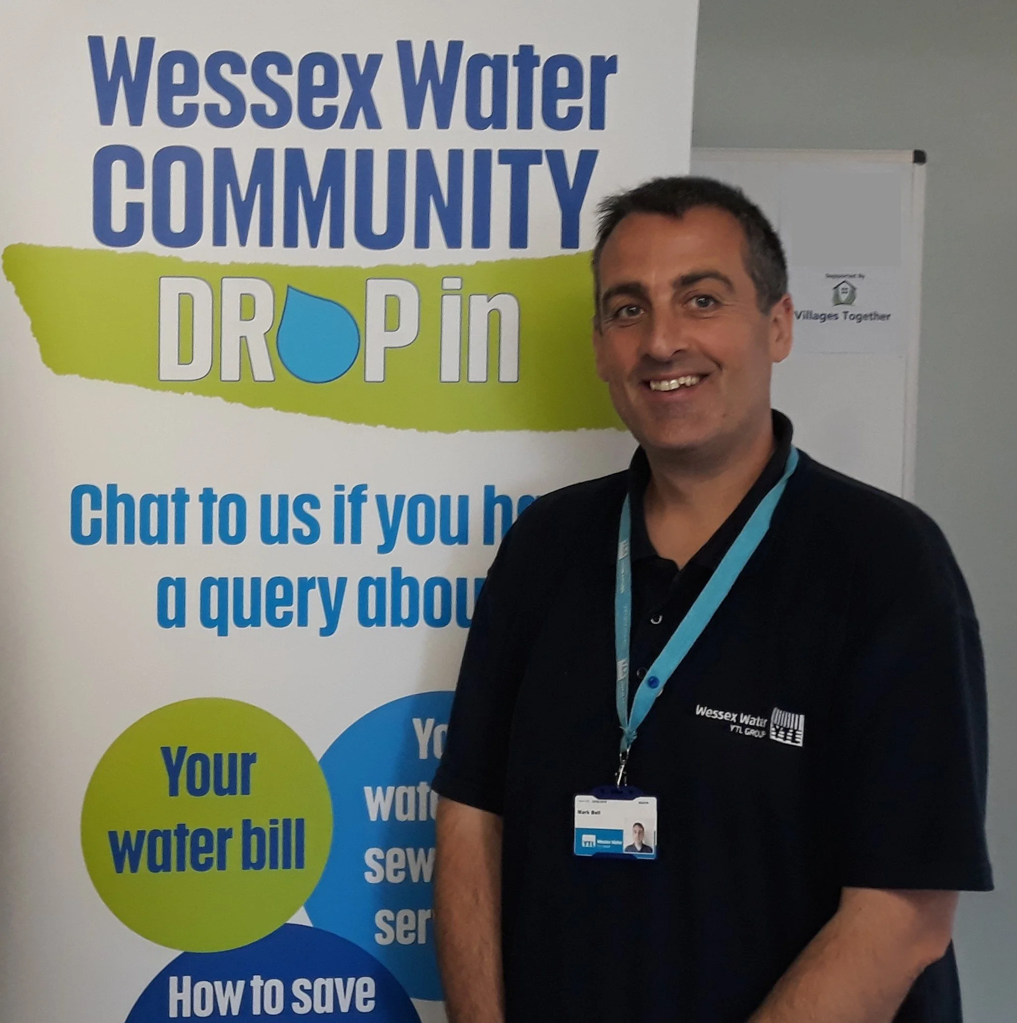 A man standing in front of a banner for Wessex Water Community Drop In event, wearing a black polo shirt with the Wessex Water logo and a name badge, smiling at the camera.