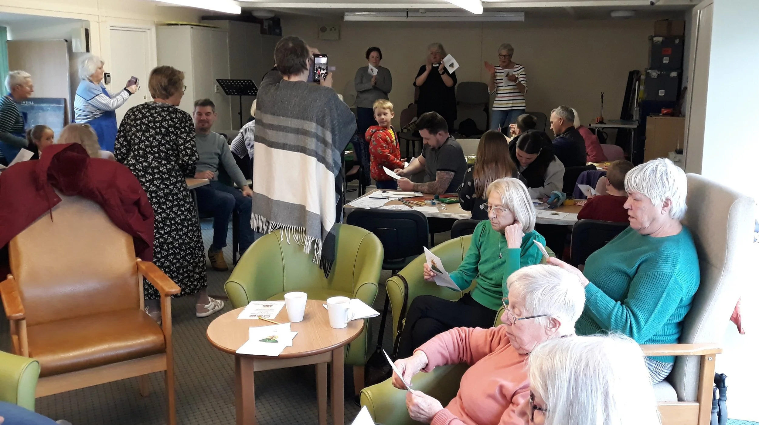 A group of elderly and middle-aged people gathered in a community room, sitting in chairs and at tables, reading papers and engaging in activities, with some standing and taking photos, in a casual social setting.