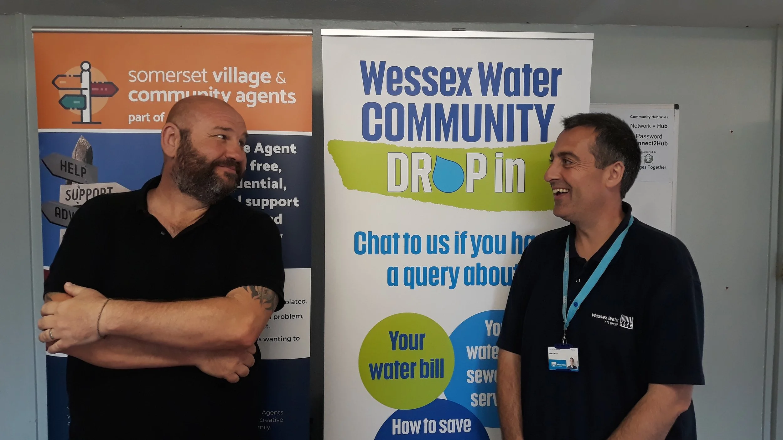 Two men standing in front of informational banners, smiling and engaging in conversation. One is bald with a beard, wearing a black shirt, and the other has short dark hair, wearing a dark shirt with a Wessex Water badge and lanyard.