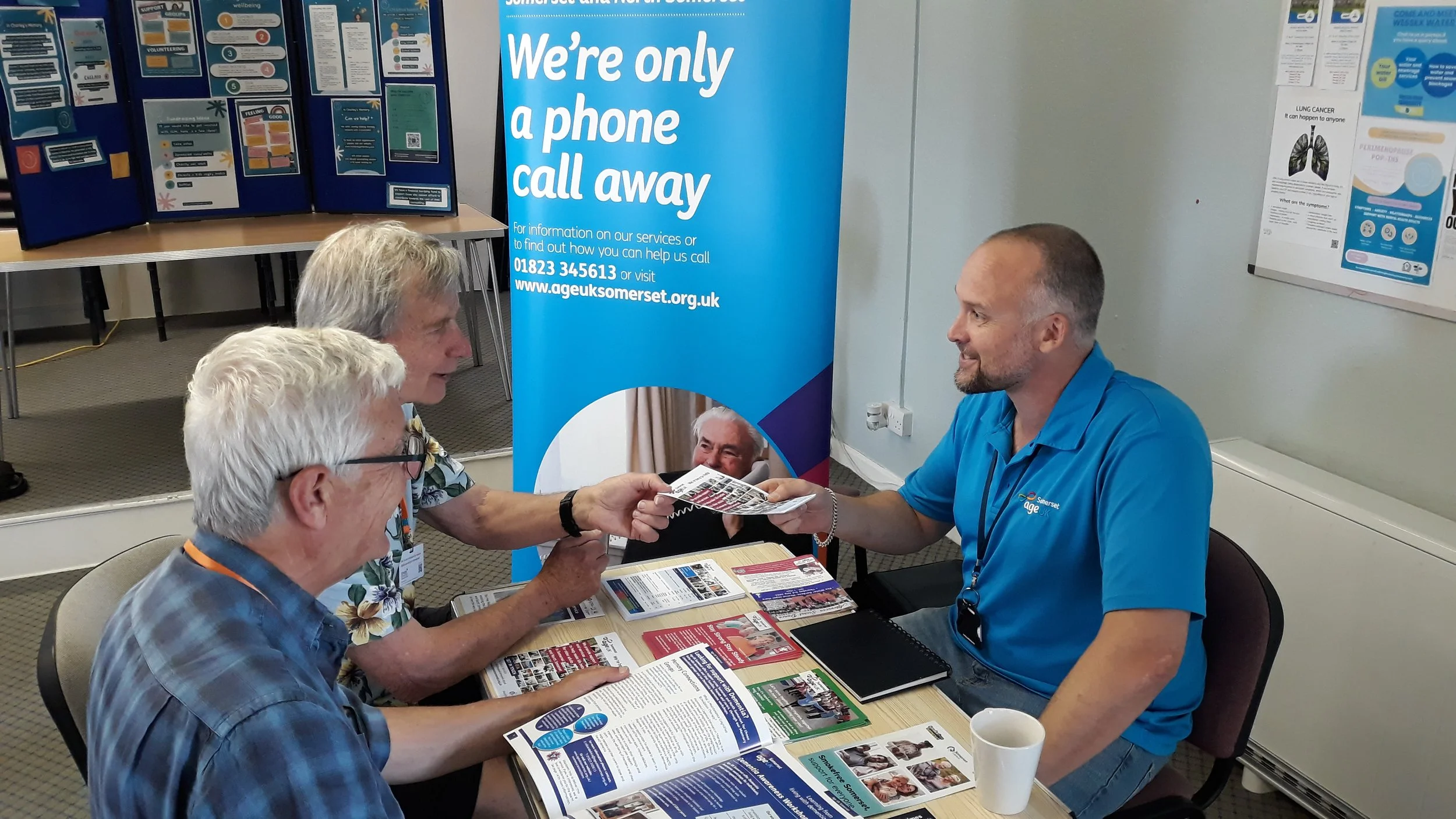 Three men sitting at a table, with one handing a pamphlet to another in front of a blue banner. The banner displays information about a phone call service from Age UK Somerset. The table has various brochures and a white mug. In the background, there are informational posters and display boards.