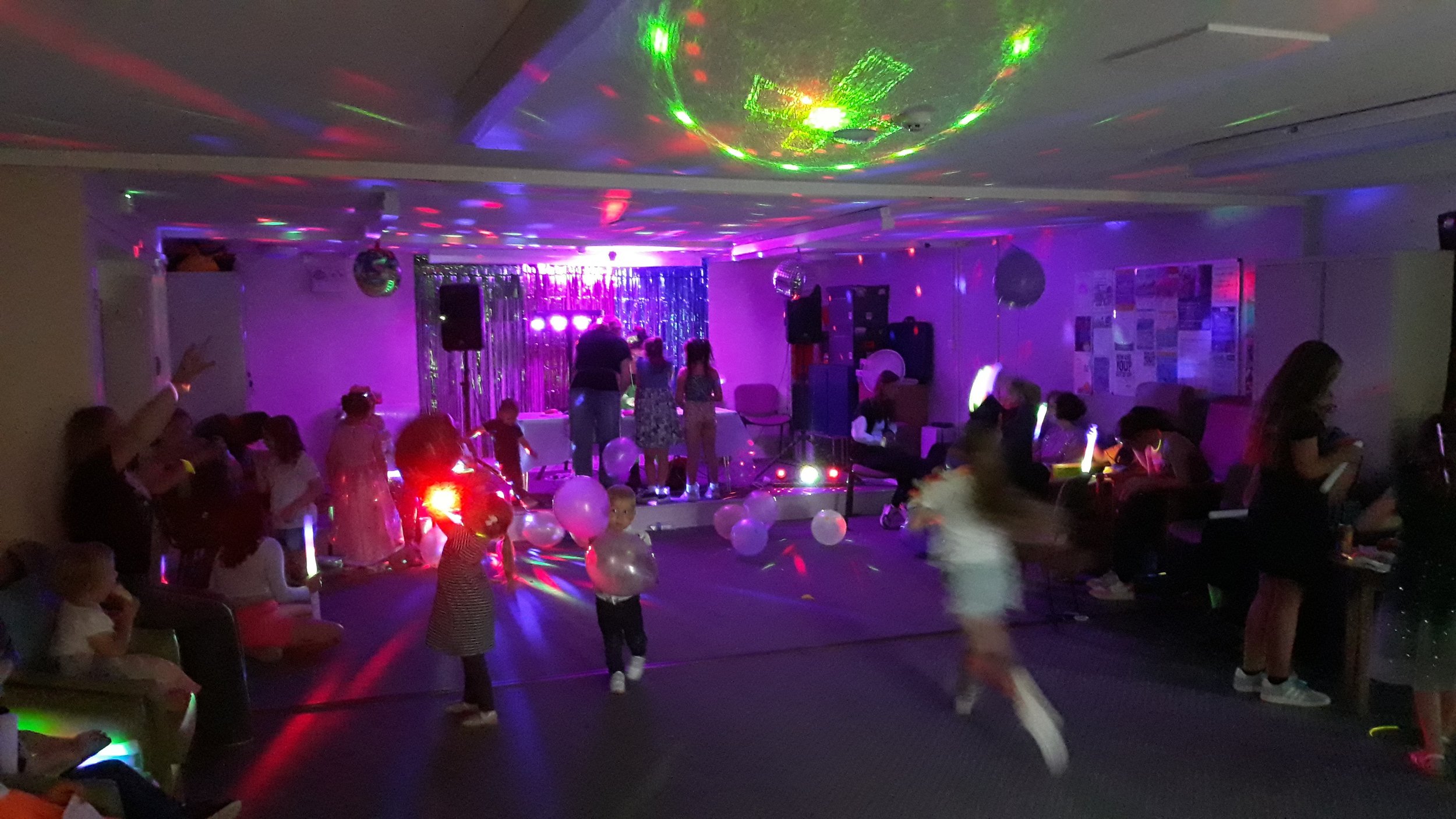 Children dancing, playing, and sitting in a dimly lit party room decorated with balloons and colorful disco lights, with a small stage area at the front.