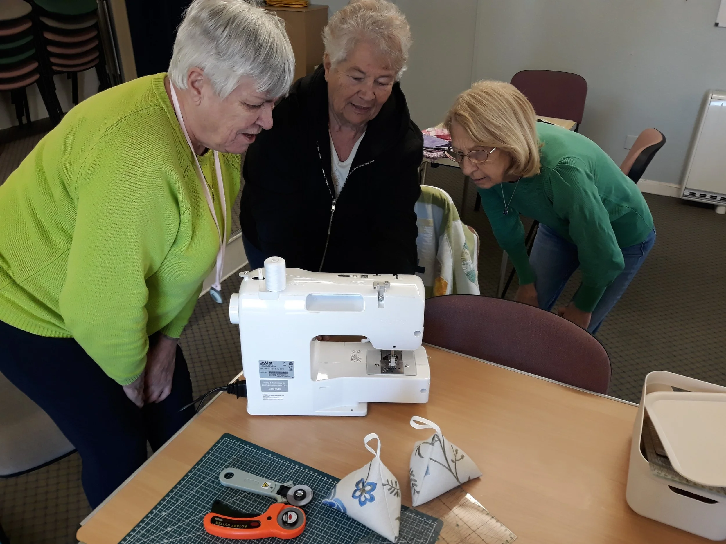 Three women gathered around a sewing machine on a table, inspecting its workings while a green cutting mat and sewing tools are nearby.