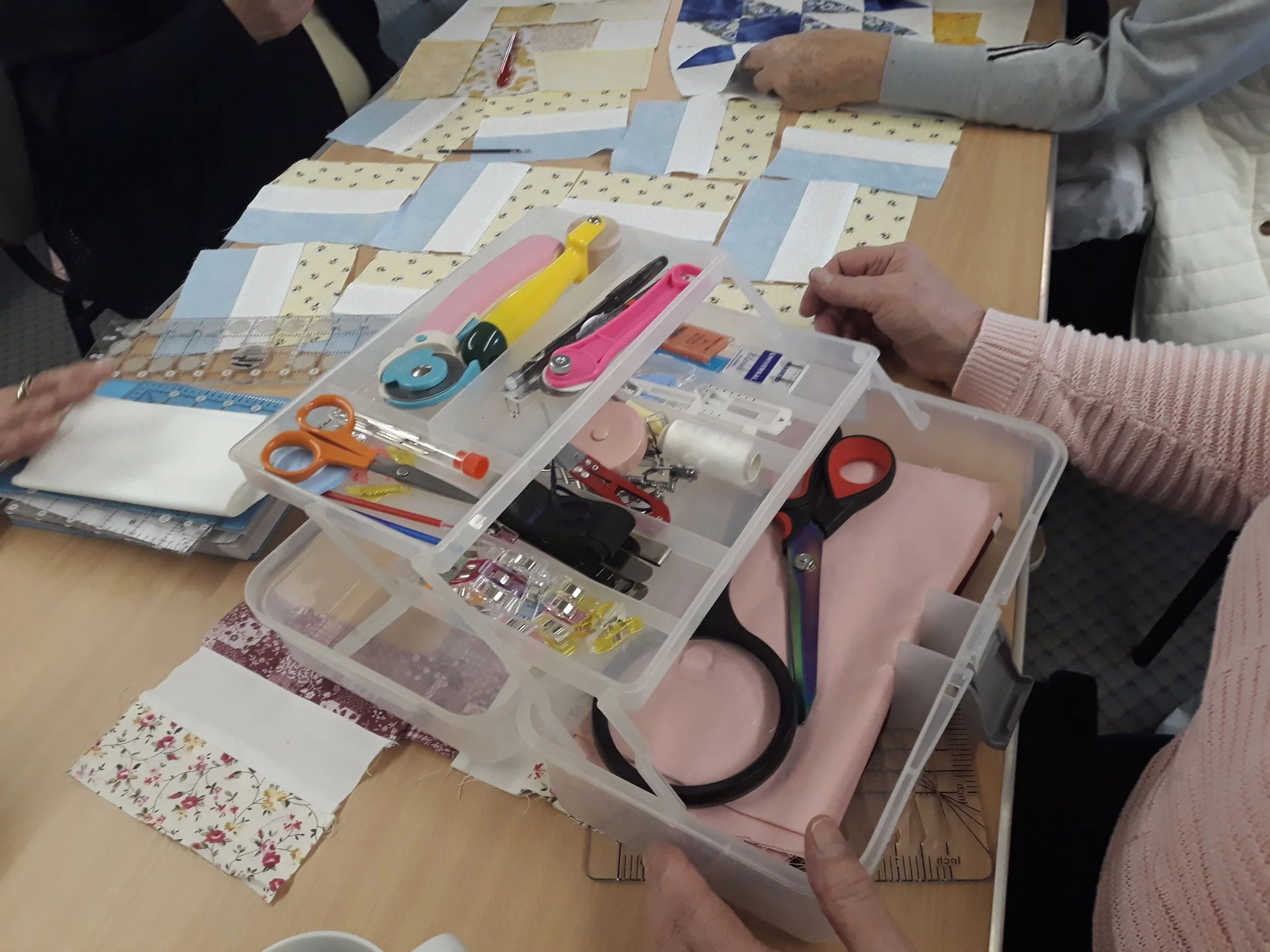 People quilting with various sewing tools and fabric pieces on a table.