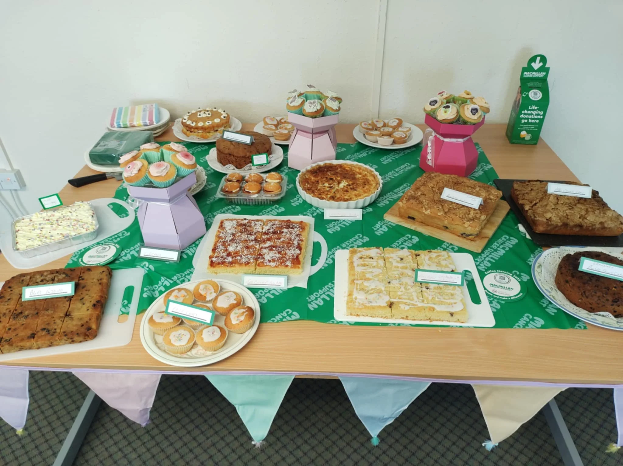 Two elderly women wearing Santa hats smiling at the camera during a Christmas celebration, sitting at a decorated table with drinks and gifts.