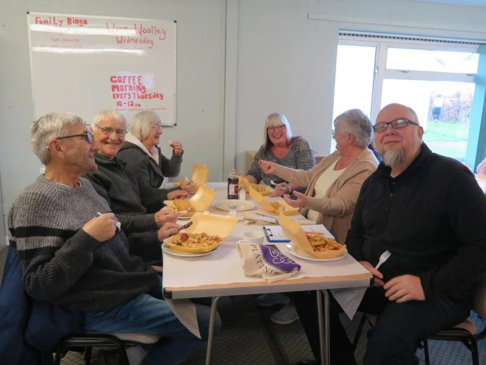 Six older adults sitting around a table, enjoying pizza and fries, with some holding utensils. They are smiling and seem to be having a cheerful gathering. There is a whiteboard in the background with handwritten events, and a window letting in natur