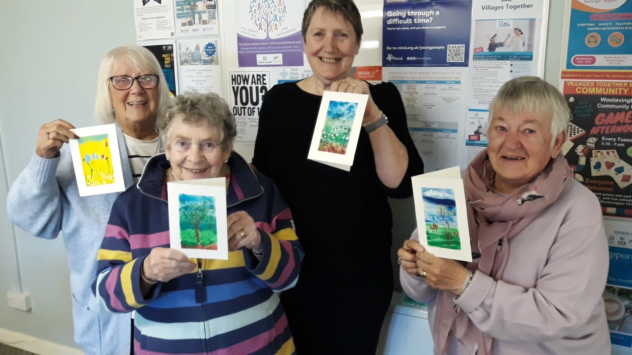 Four elderly women smiling and holding colorful drawings of trees and landscapes in an indoor community space, with posters and flyers on the wall behind them.