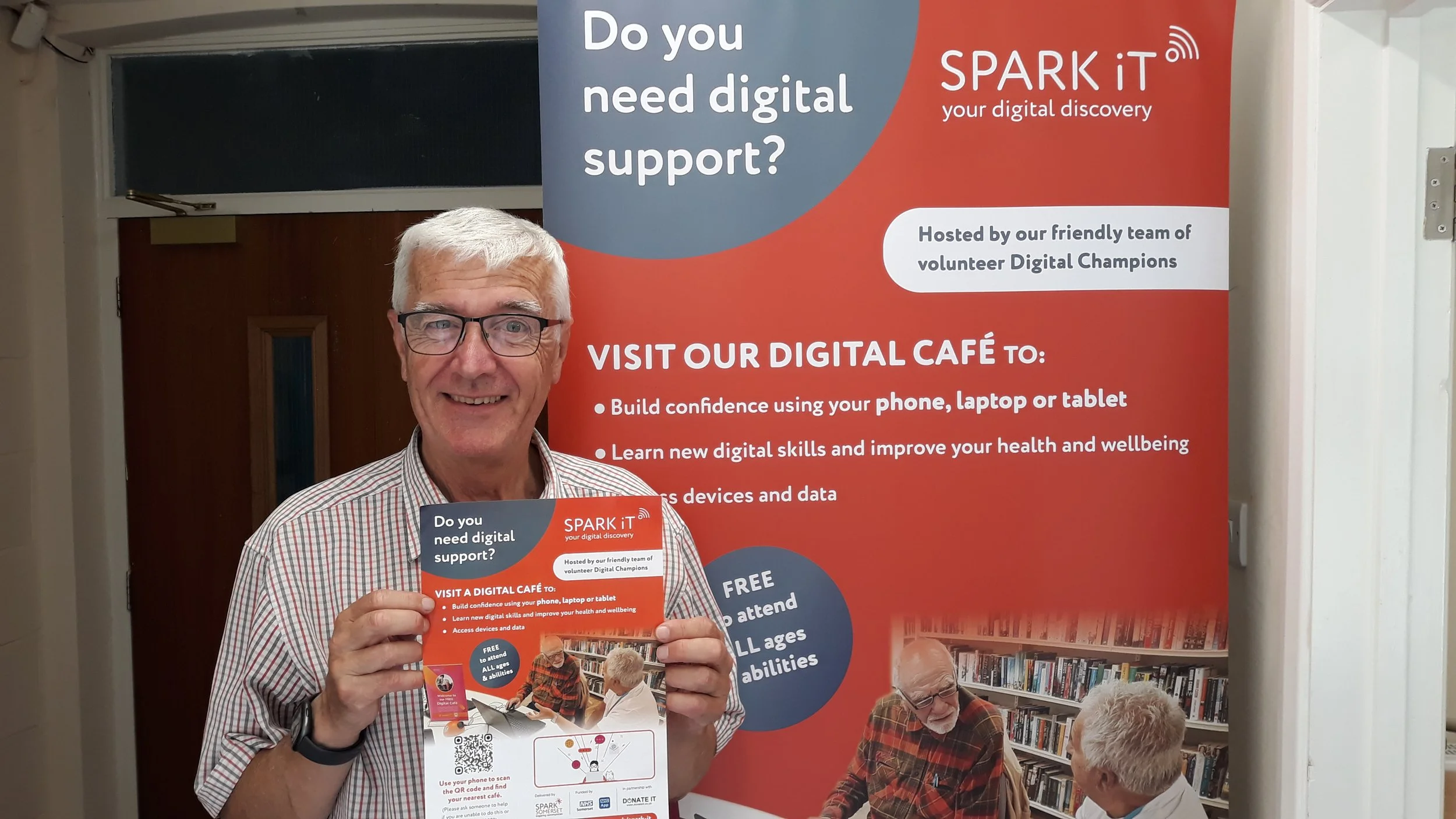 An elderly man wearing glasses and a plaid shirt holding a flyer in front of a large promotional poster for a digital support program called SPARK IT, which offers digital skills and confidence building.