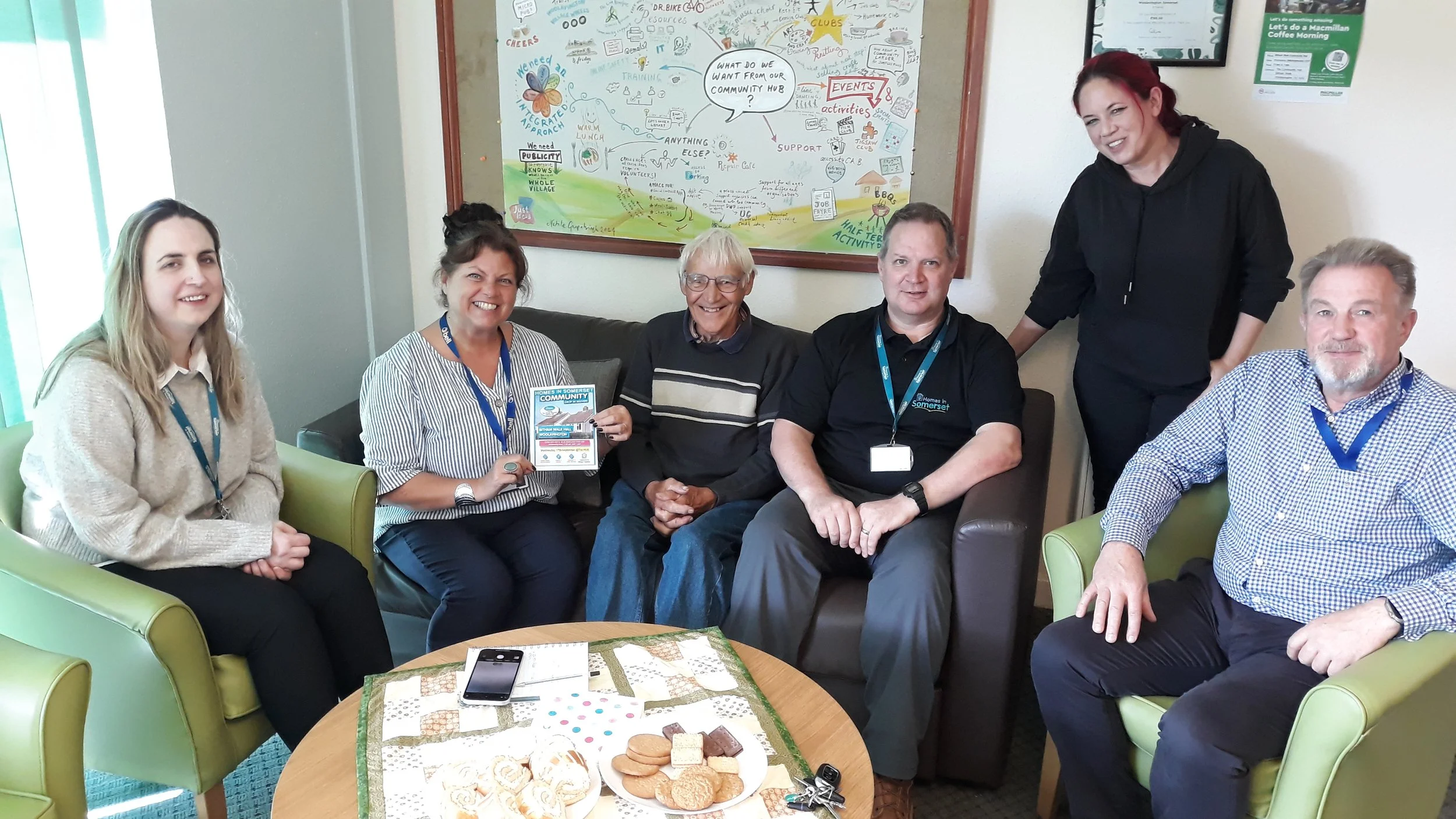 Group of six people sitting and standing in a casual meeting room, with a table of cookies and a smartphone in the foreground. They are smiling and looking at the camera, and one person is holding a flyer.