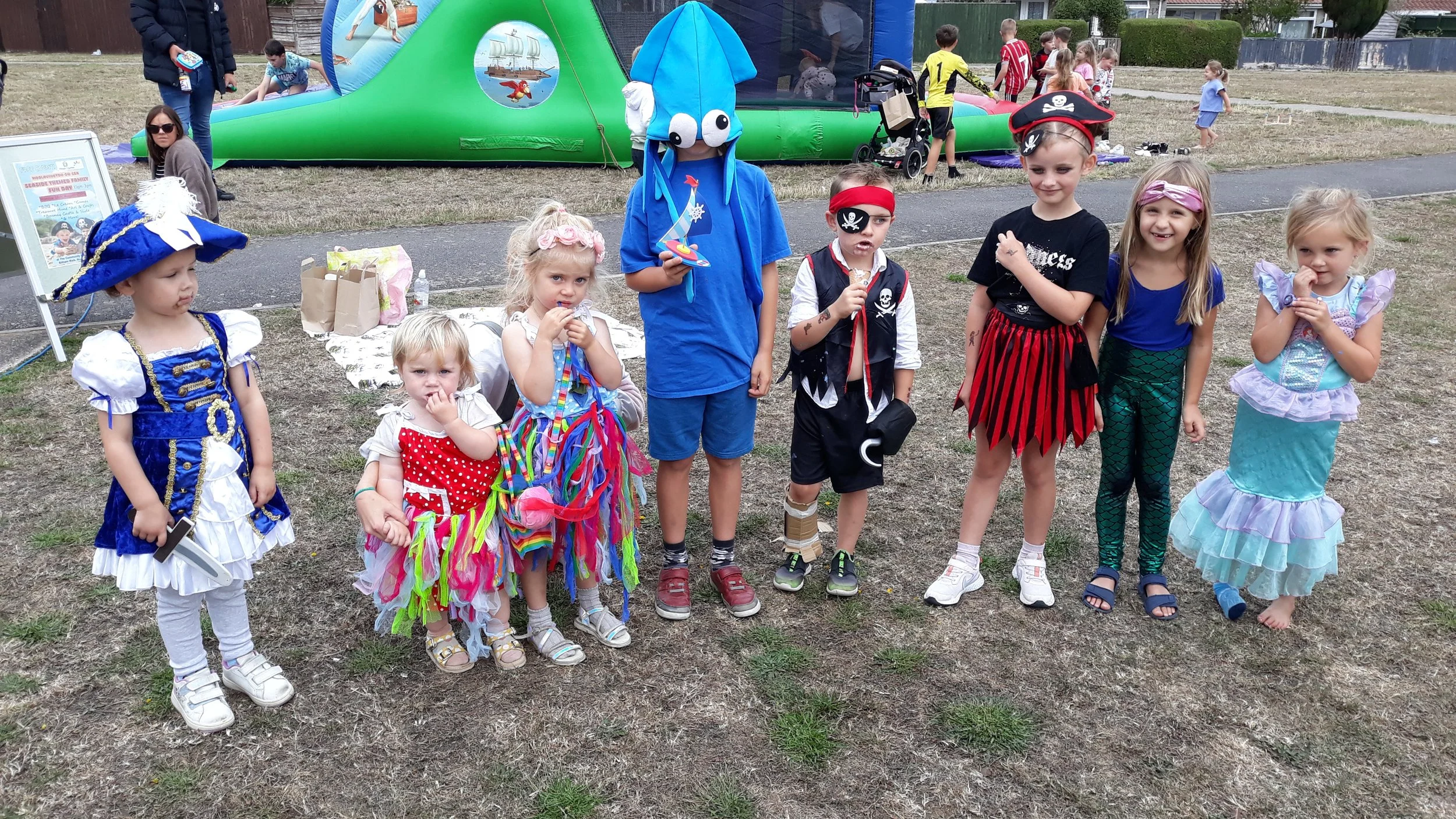 Group of children dressed in various costumes, standing outdoors on a patchy grass field during a festive event, with a bouncy castle and adults in the background.