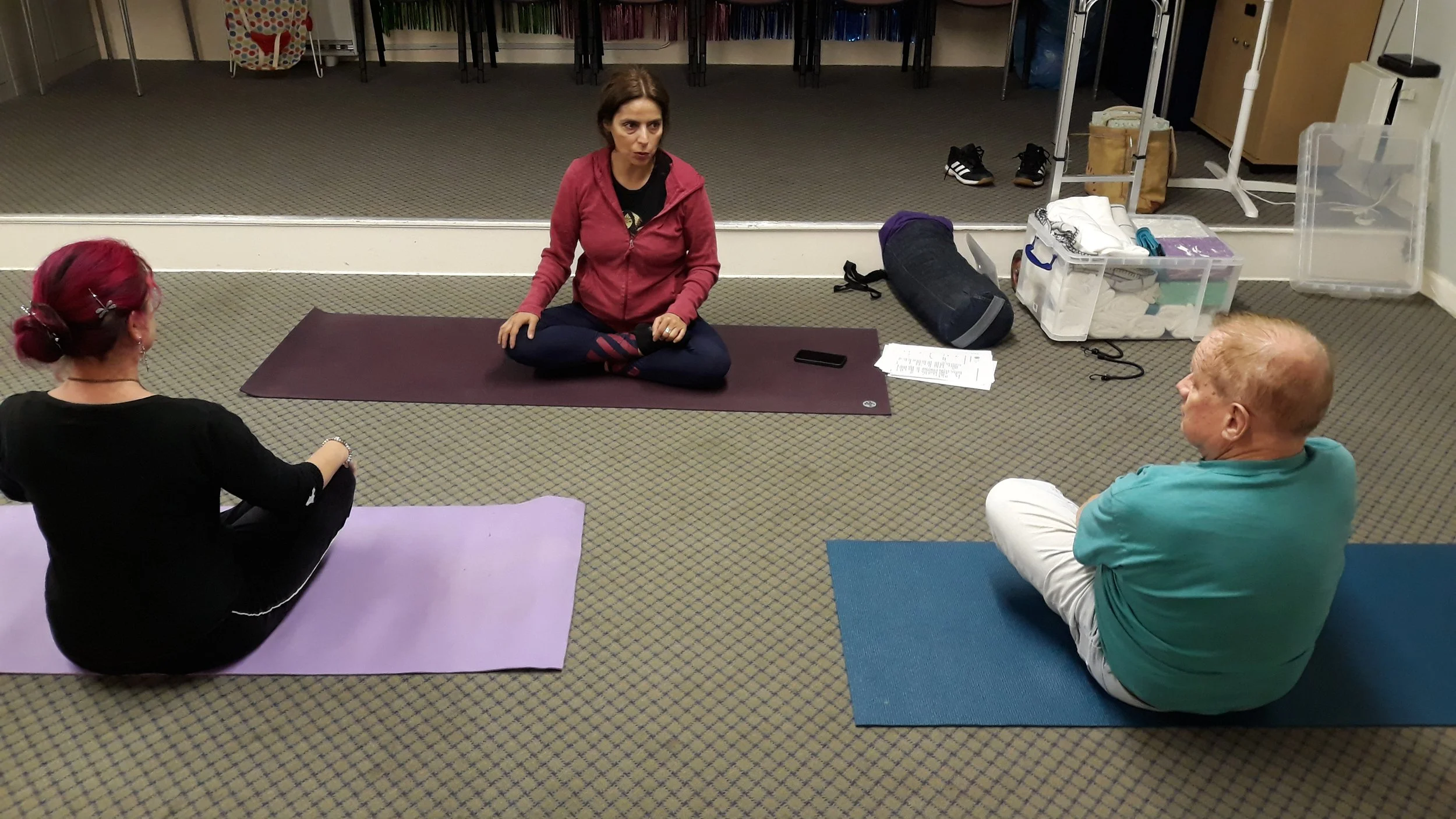 Three people sitting on yoga mats in a room, engaged in a meditation or yoga class. One woman with red hair and a black outfit is on a purple mat, another woman in a pink hoodie is sitting cross-legged on a maroon mat, and an older man with white hai