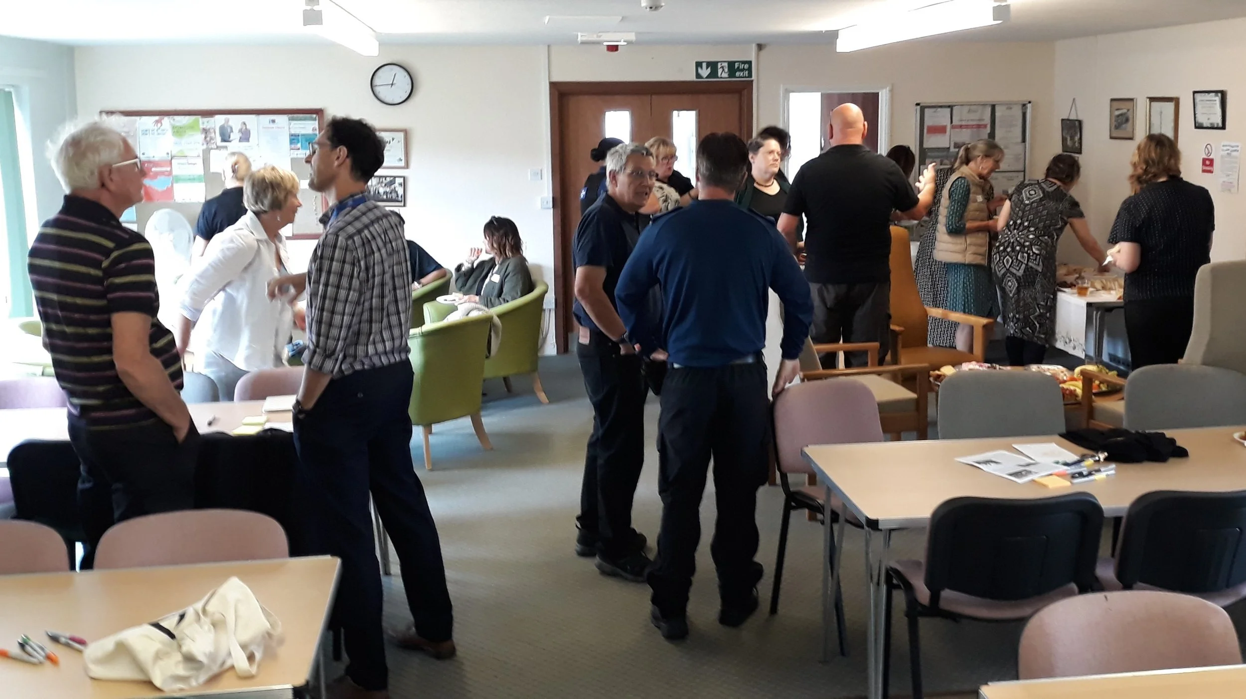 People gathering in a community room with tables, chairs, and a food table, some engaged in conversation and others serving or preparing food.