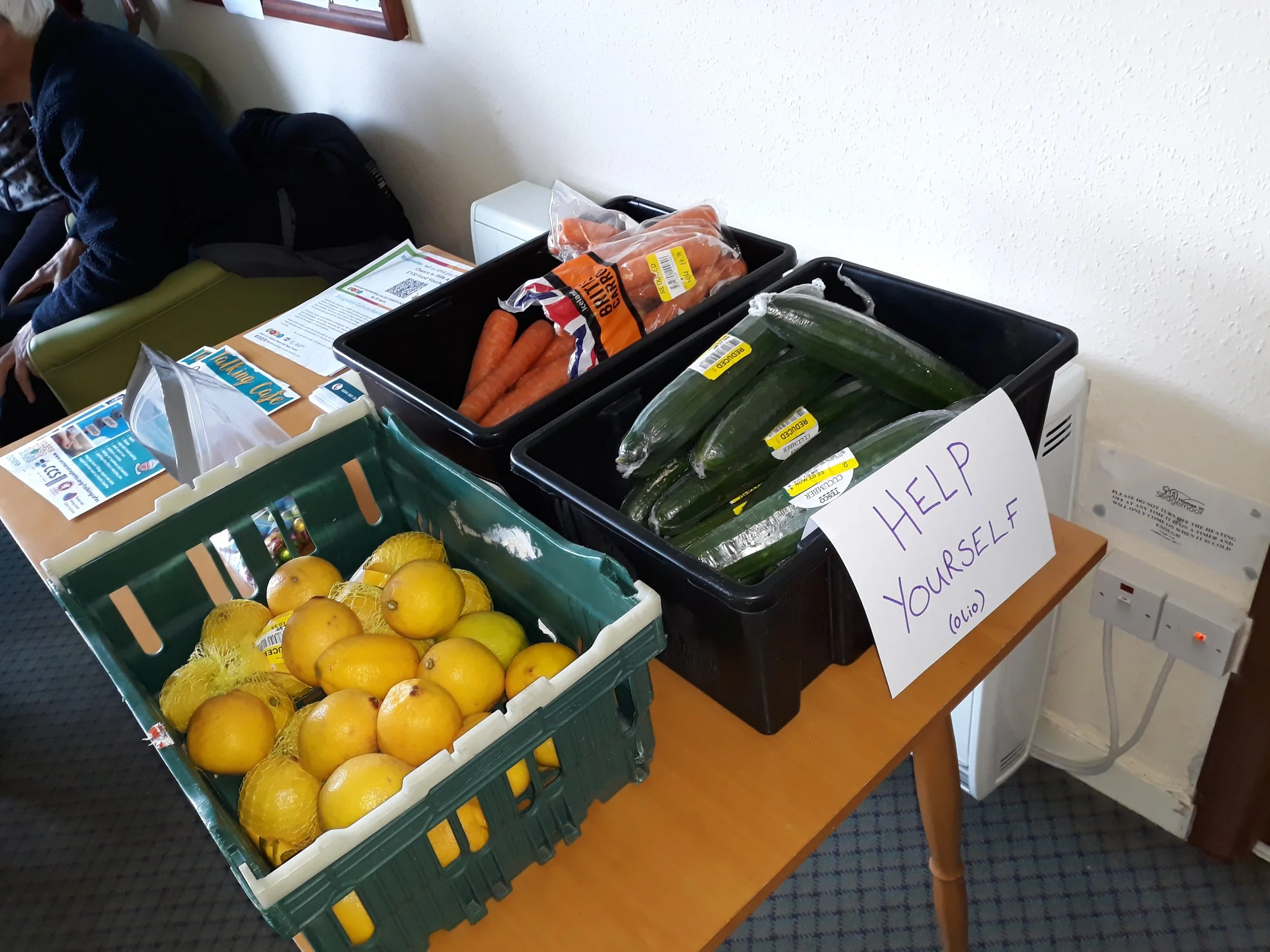 Table with crates of fresh vegetables and fruit, including lemons, carrots, and zucchini, with a handwritten sign saying 'Help Yourself'.