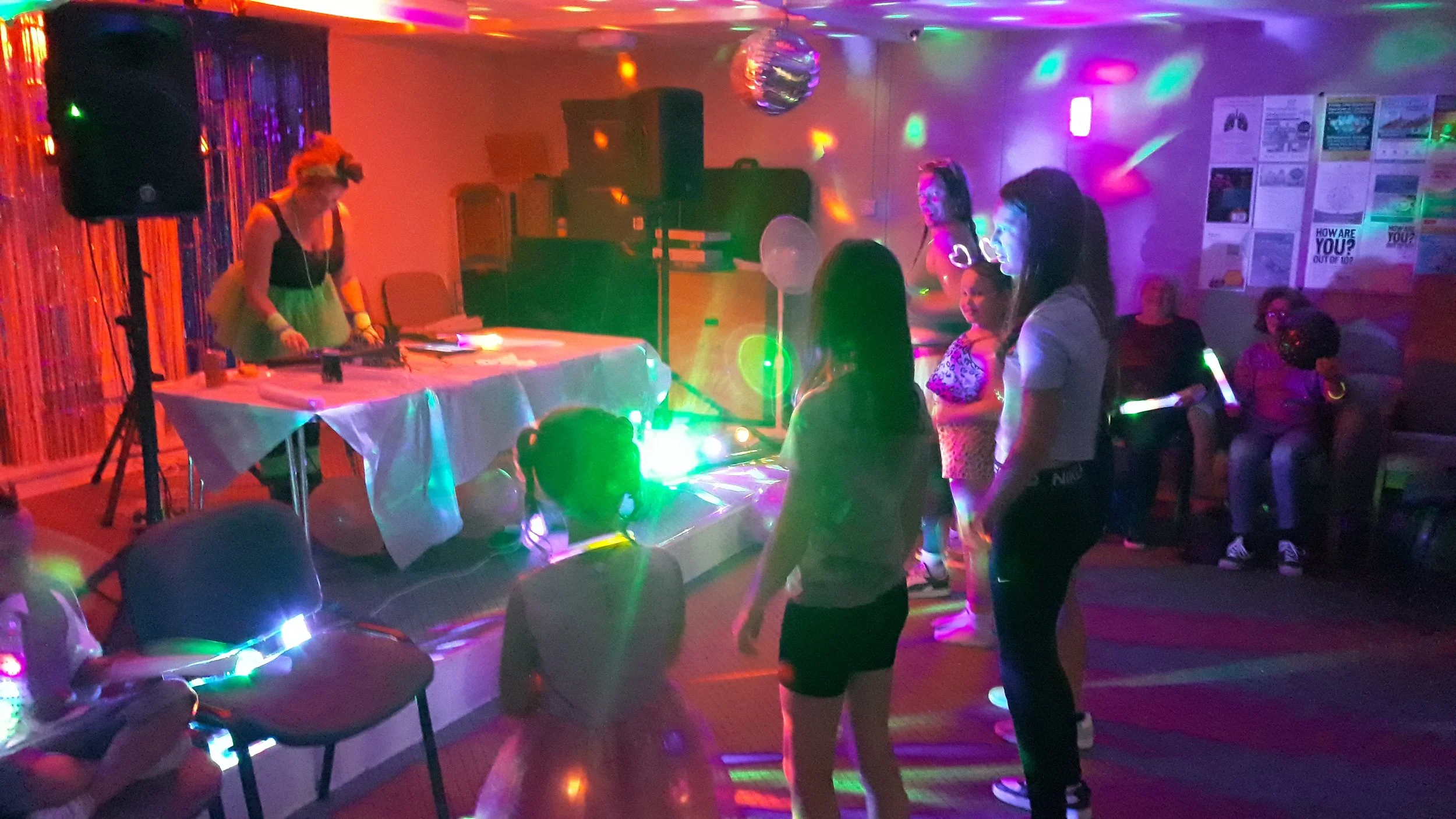Children and adults dancing and socializing at a party with disco lighting and a DJ, in what appears to be a community center or hall.