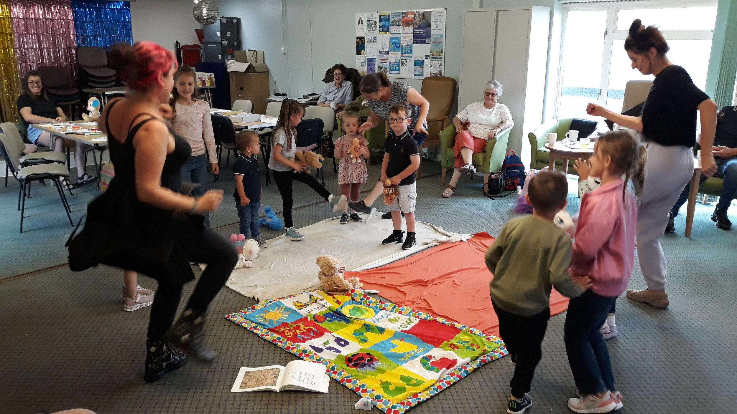 Children and adults participating in a group activity in a community room, with toys, books, and colorful decorations.